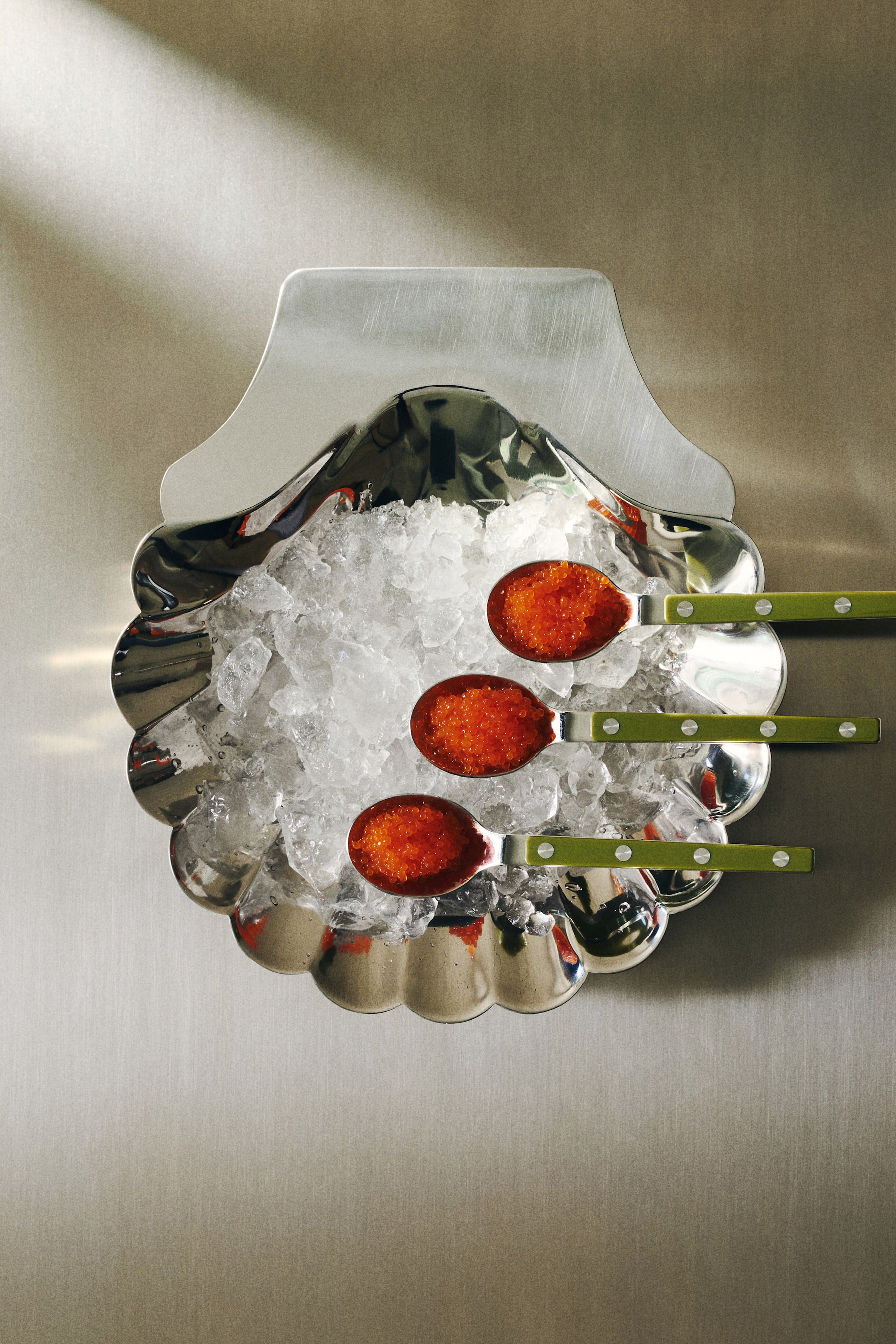 Agrandir l'image: Un bol de service en acier inoxydable poli présente une forme de coquillage vue de dessus, avec un bord festonné sur sa moitié inférieure et une section supérieure plate. Le bol est rempli de glace pilée, et trois cuillères contenant du caviar orange reposent sur la glace.