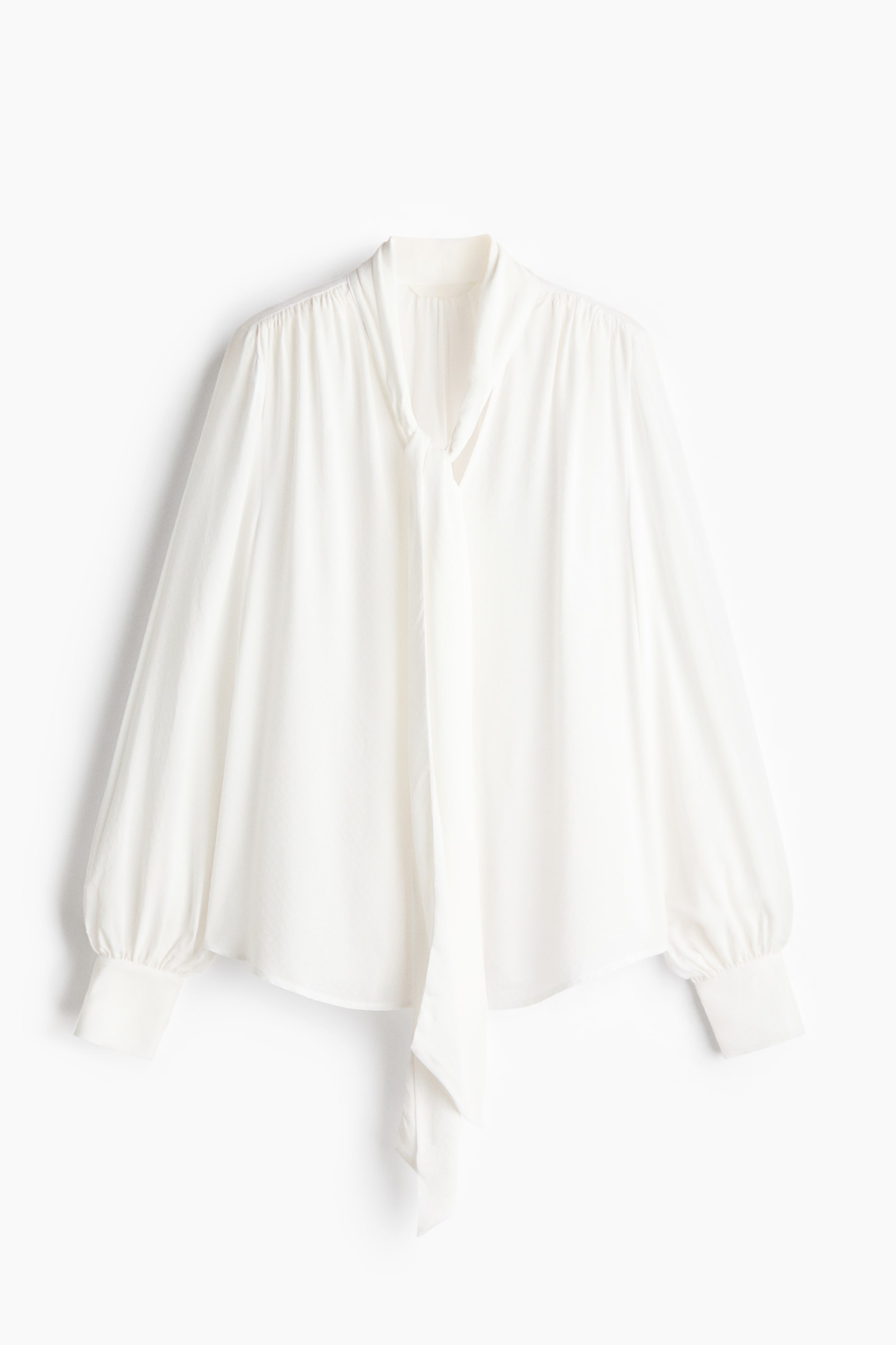 Ver imagen más grande: Una blusa blanca se presenta de frente, con cuello alto y un largo lazo, mangas abullonadas recogidas en los hombros y puños anchos.