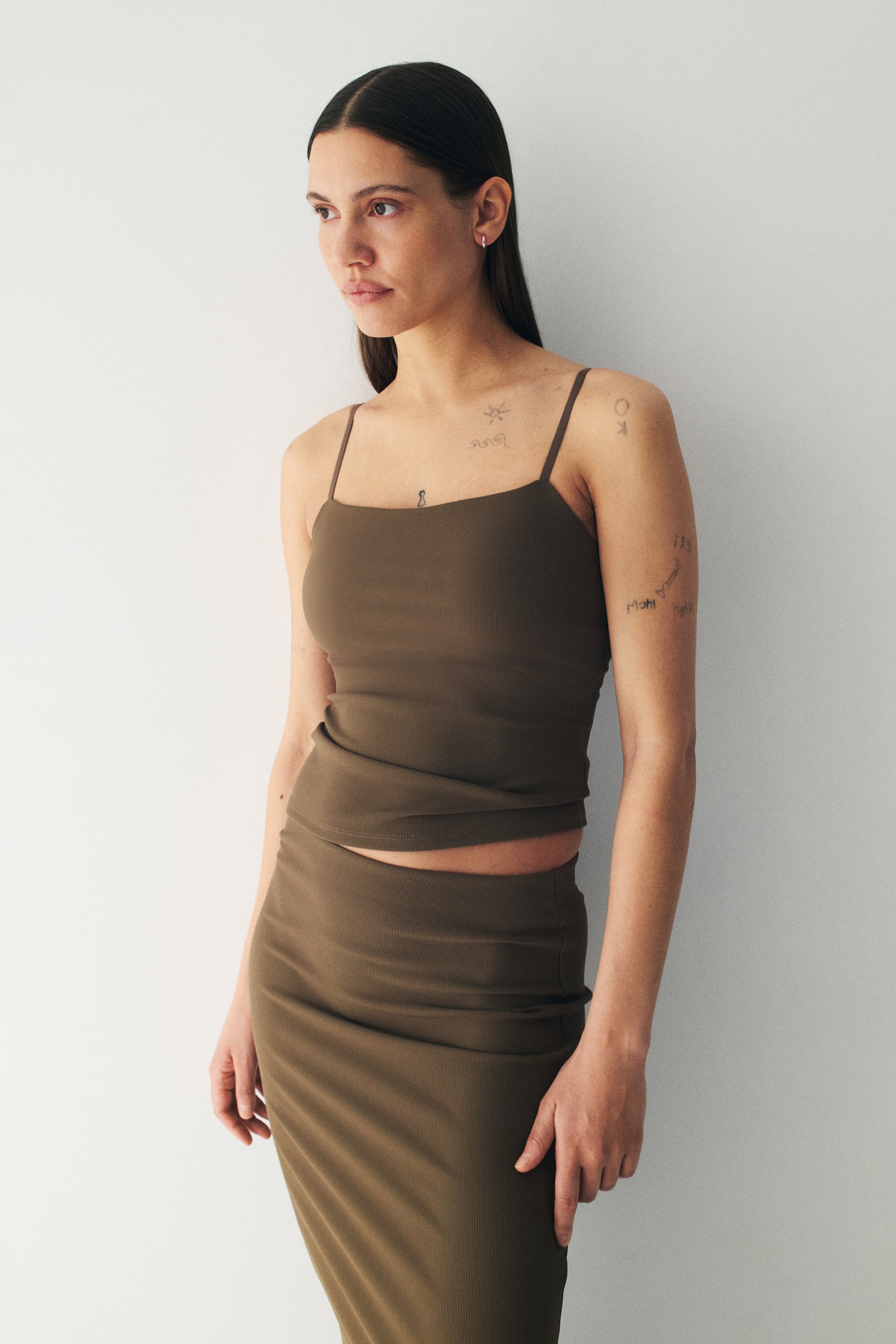 View larger image: A dark olive green ribbed camisole top and a matching ribbed midi skirt are being presented from a slight front-left angle. The camisole is featuring thin spaghetti straps and a square neckline, while the form-fitting skirt is extending below the knee.