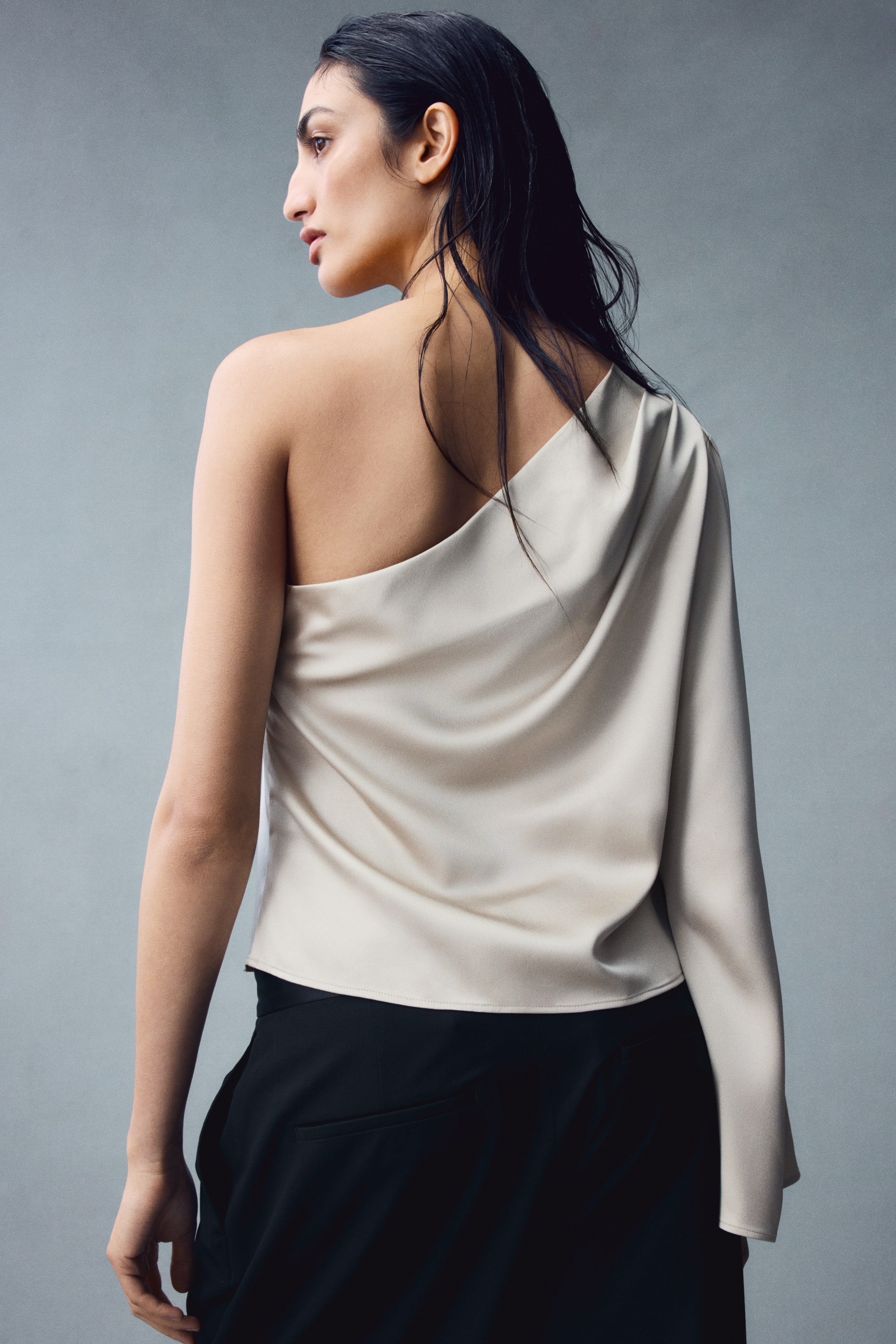 View larger image: A light beige, one-shoulder top in draped satin is being worn by the model, featuring an asymmetrical neckline and a single long, wide sleeve. The top is styled with black trousers, showcasing the draped fabric across the back.