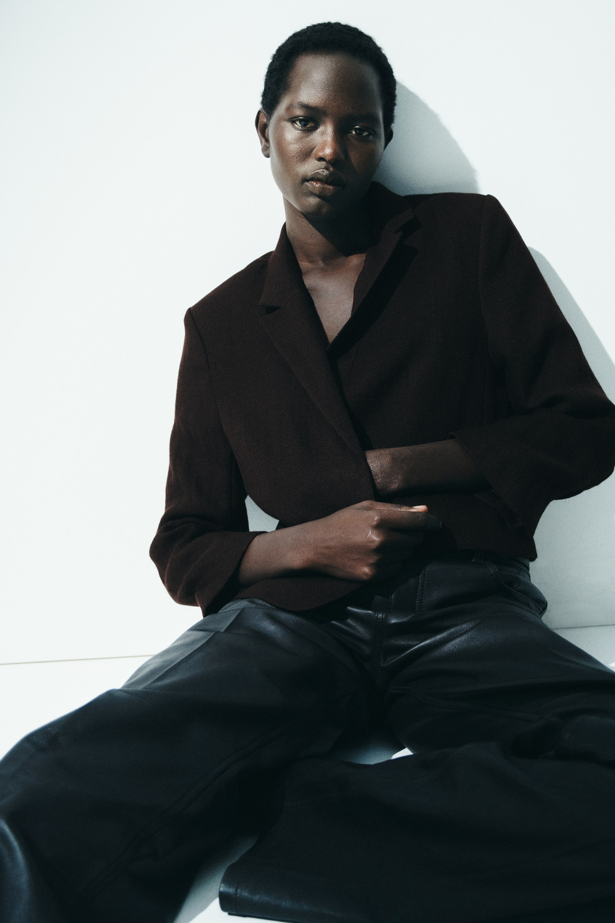 View larger image: Dark brown blazer with notch lapels, showcasing a relaxed, cropped fit. Layered over a dark inner garment and matched with black faux leather wide-leg trousers, which feature prominent front seams.