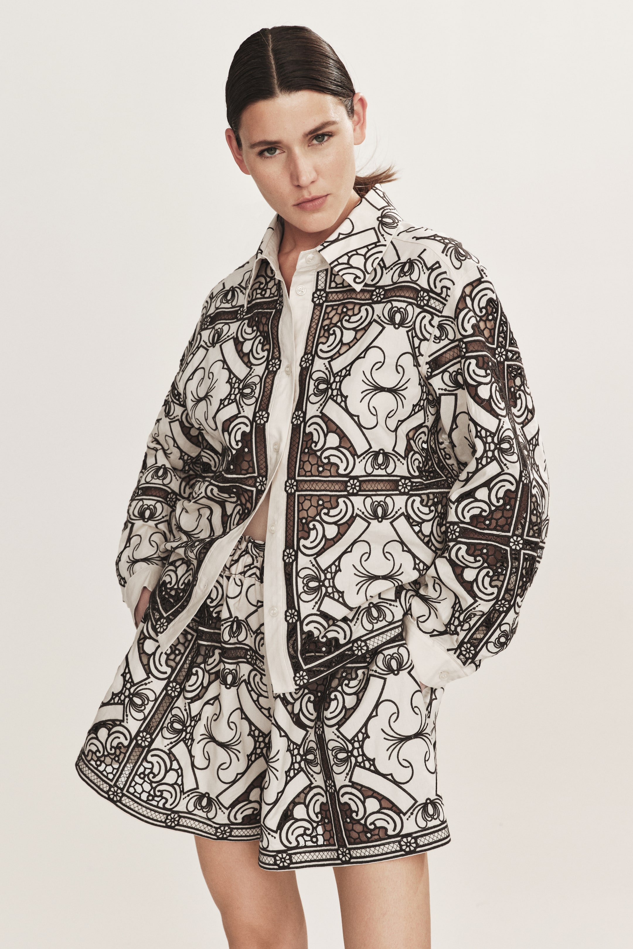 Ampliar la imagen: El conjunto de camisa de cuello grande y shorts a juego está orientado hacia adelante, presentando un intrincado patrón en negro y blanco roto con motivos geométricos y florales, incorporando detalles transparentes similares al encaje. La camisa está diseñada con mangas largas y cierre de botones al frente, y los shorts de corte holgado llegan a la mitad del muslo.