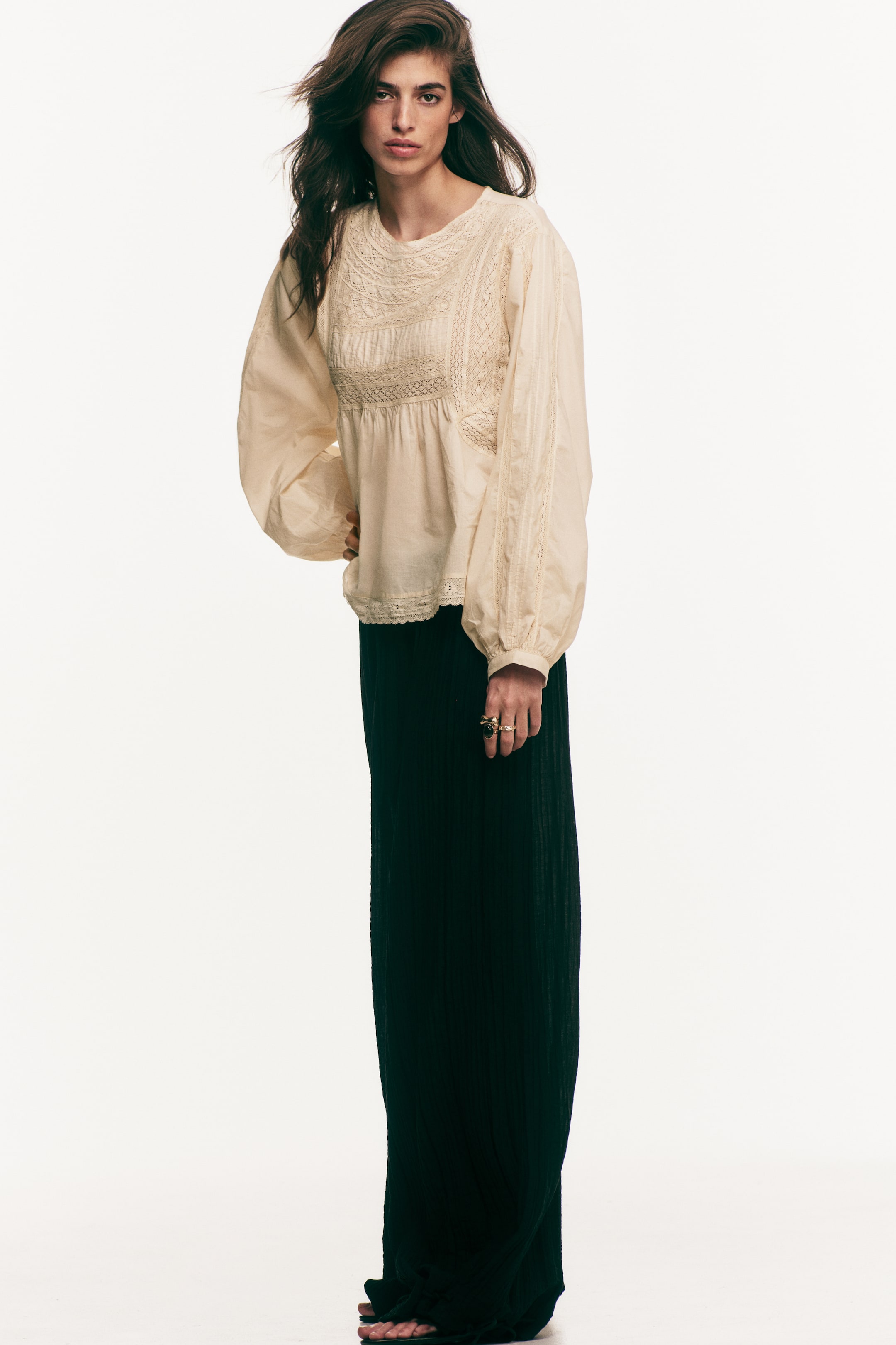 View larger image: A cream-coloured cotton weave blouse, featuring a round neckline, a lace yoke with pintucked details, and lace trims on the long balloon sleeves with narrow cuffs, is being worn by the model. A gathered seam is visible under the bust, and the blouse is styled with a dark skirt while the model is standing with one hand on her hip.