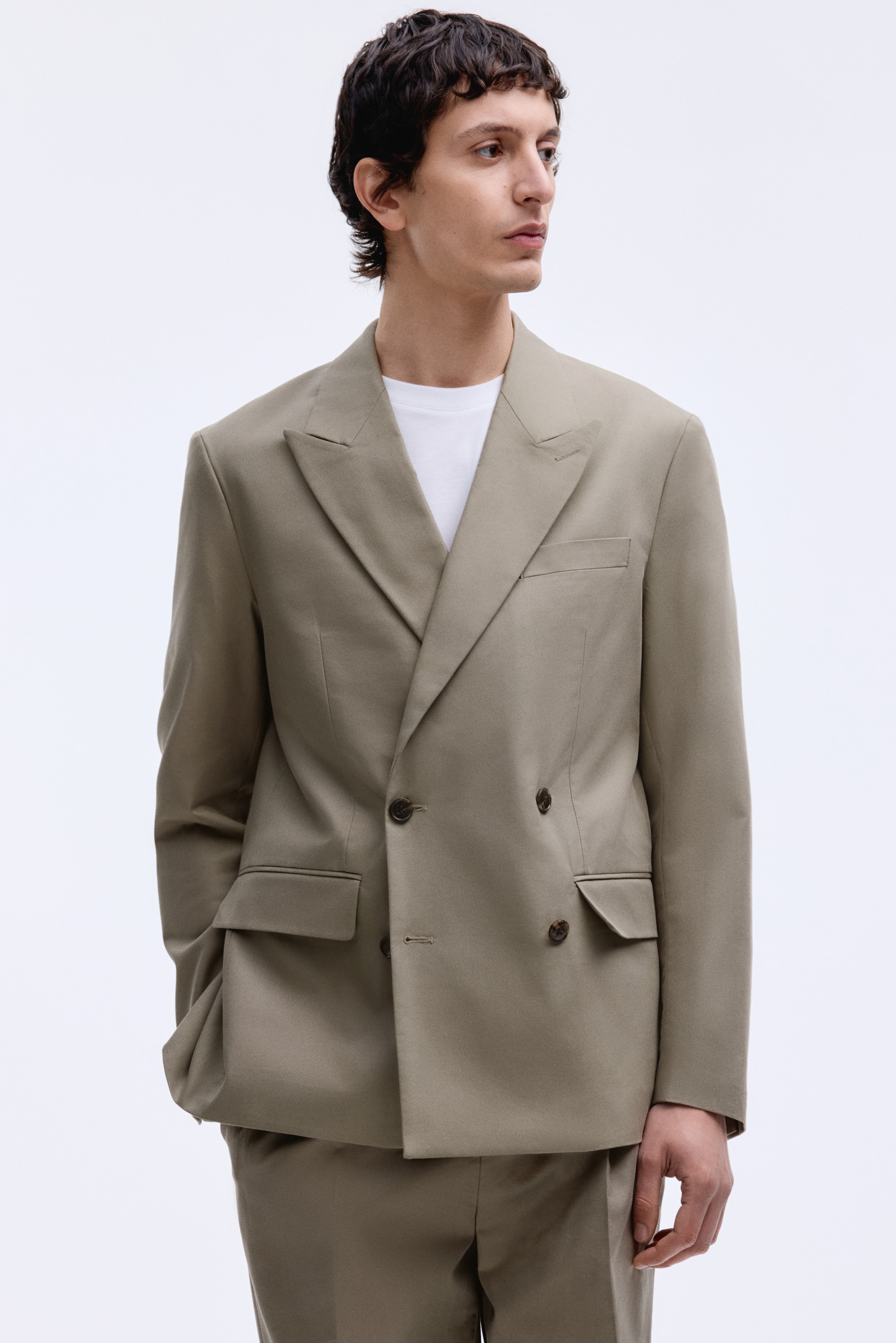 Agrandir l'image: Un blazer croisé kaki en tissage doux, avec des revers en pointe, une poche poitrine et des poches avant passepoilées avec rabat, est porté par le mannequin. Le mannequin porte le blazer à coupe ample sur un t-shirt blanc et un pantalon assorti, avec une main reposant dans une poche avant.