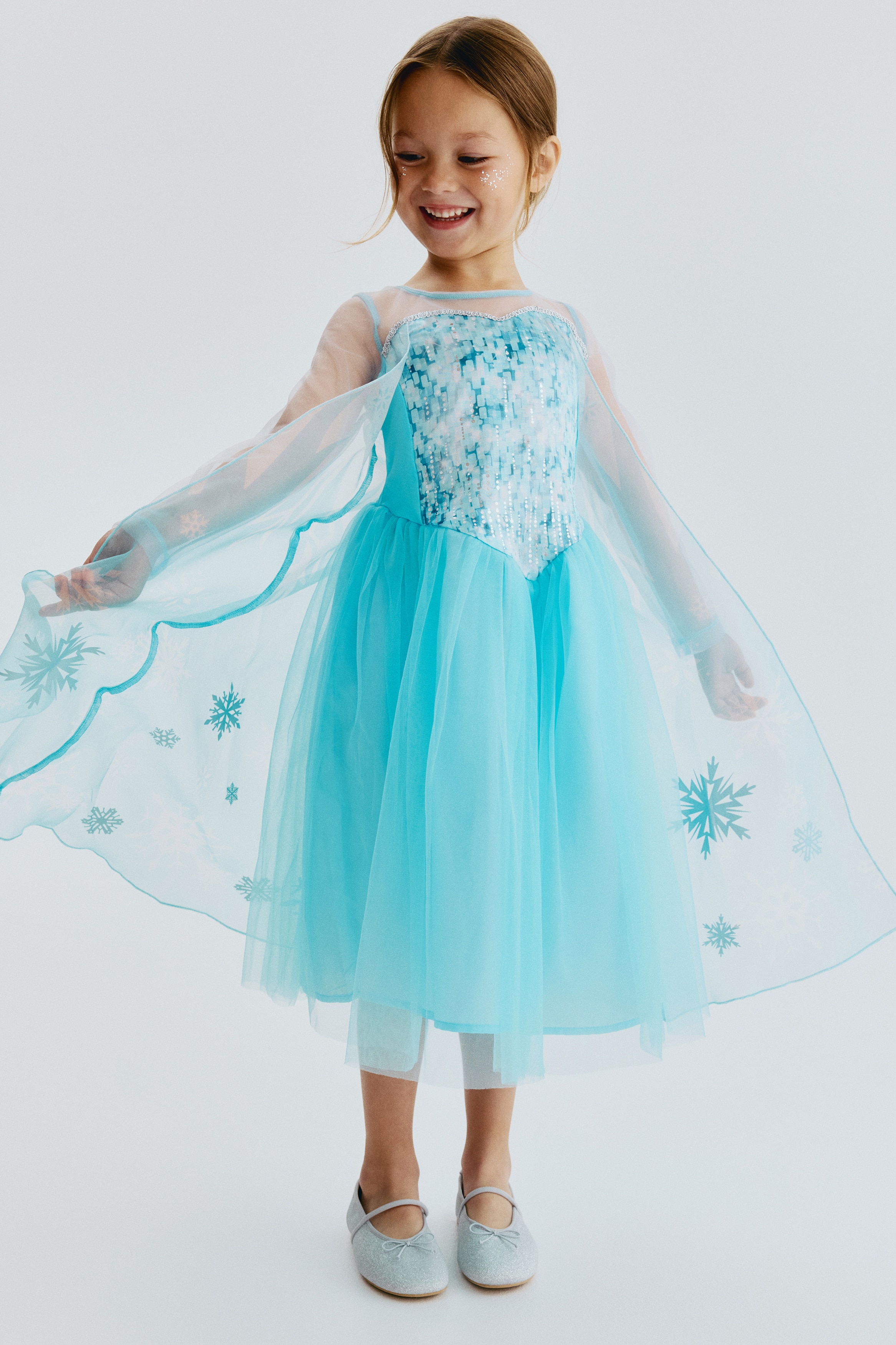 A light blue dress is showcasing its front, featuring a sheer round neckline and elbow-length sleeves. The bodice displays a heart-shaped panel with a glittery light blue and silver pattern, outlined in silver, and the skirt presents a sheer overlay adorned with silver glitter and snowflake designs.