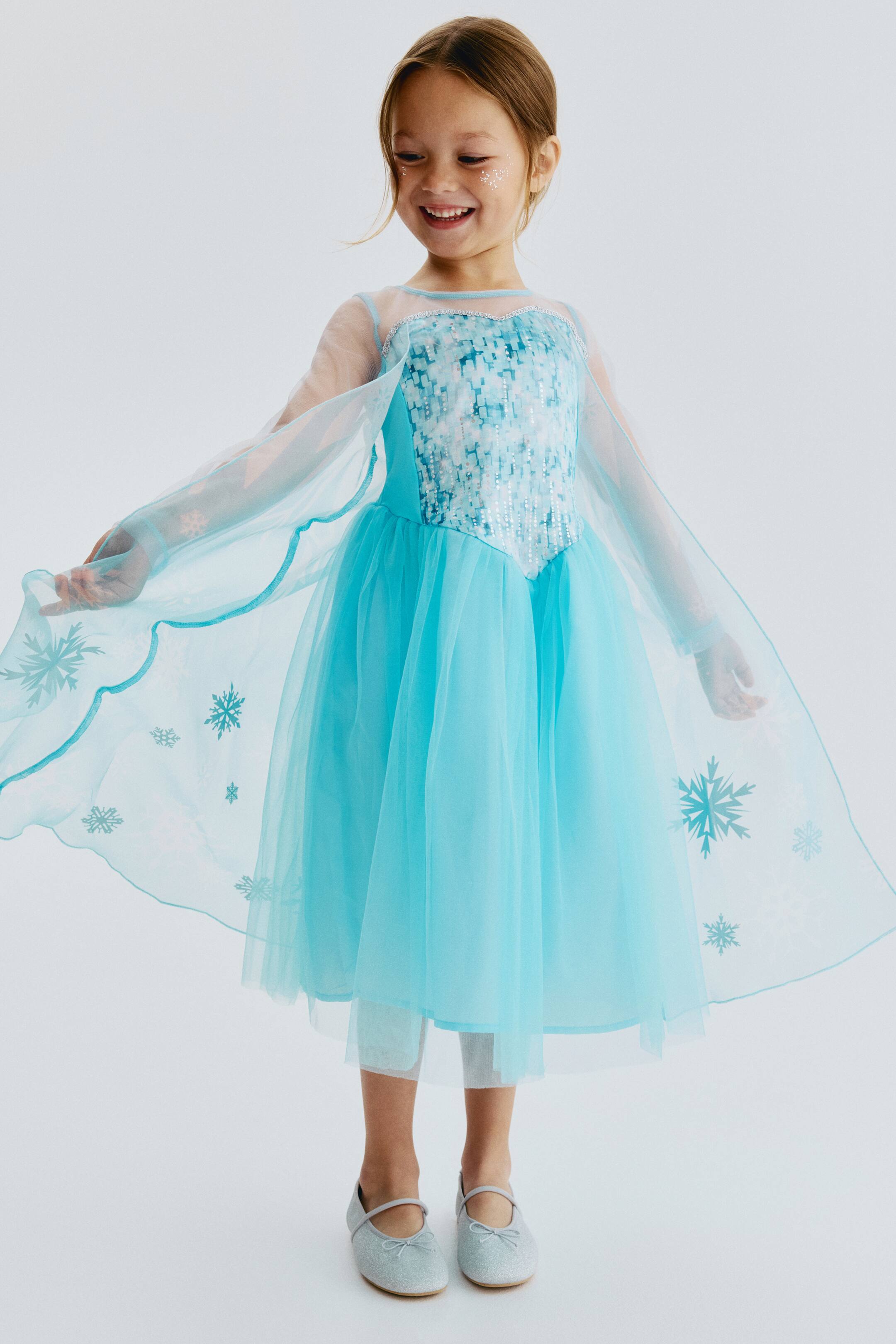 View larger image: A fancy dress costume is shown from the front, featuring a light blue satin bodice with a sequined overlay and sheer sleeves. A lined, flared mesh skirt complements the bodice, while a sheer cape with snowflake patterns flows from the shoulders.