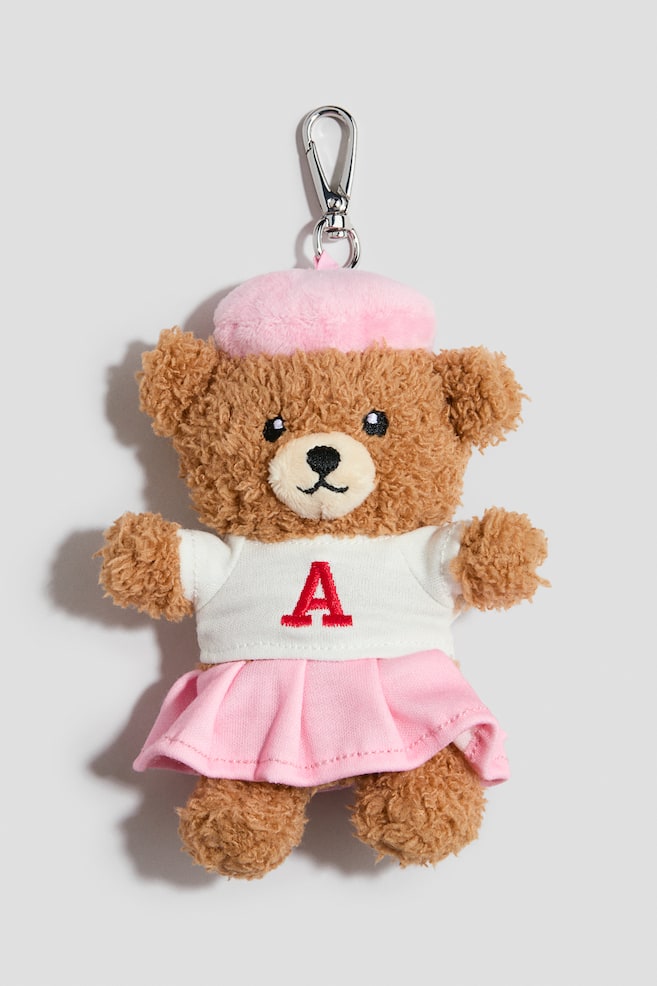 A bag charm is shown from the front, featuring a brown, textured plush teddy bear suspended from a silver-toned metal carabiner hook. The teddy bear is wearing a light pink beret, a white short-sleeved T-shirt with a red embroidered letter "A" on the chest, and a matching pink pleated skirt. - 1