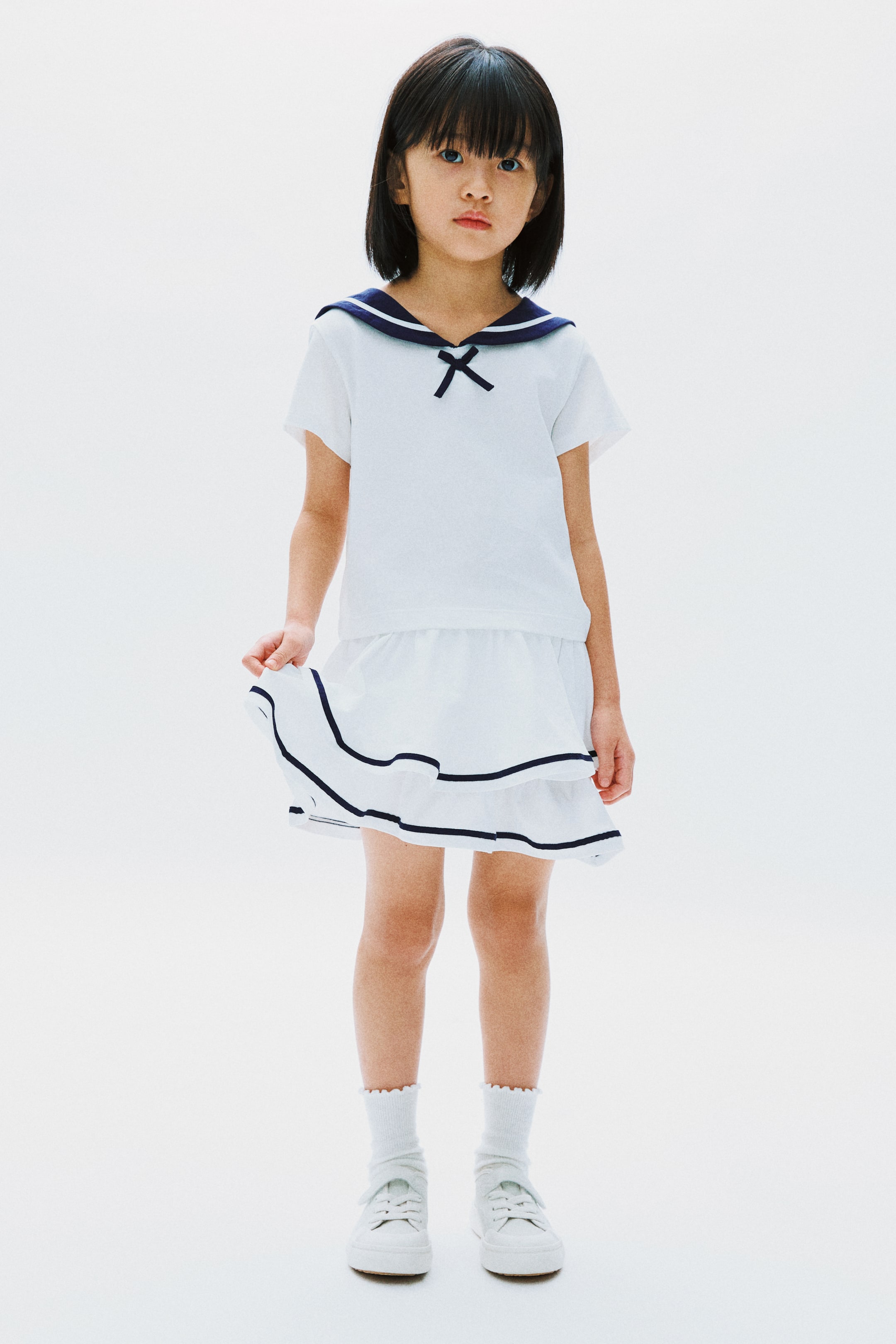 View larger image: The model is wearing a white short-sleeved top with a navy sailor collar and a small navy bow, perfectly matched with a white tiered skirt. The skirt is adorned with two navy stripes, and the model is elegantly lifting its hem.