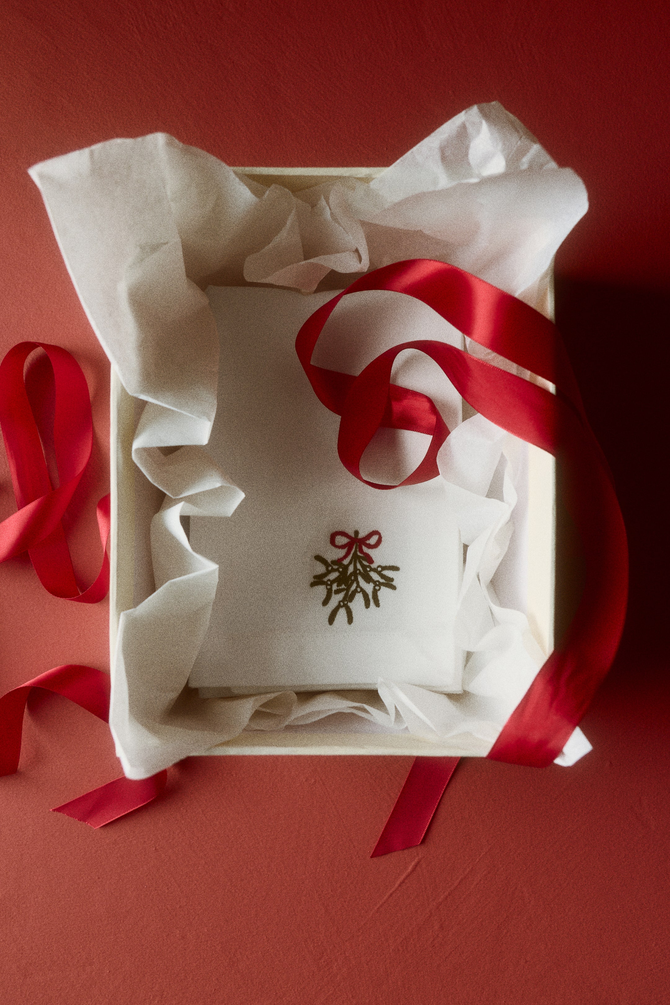 View larger image: A white cotton sateen napkin, folded to show a small embroidered mistletoe design with a red bow, is resting inside a gift box lined with white tissue paper. A red satin ribbon is draping over the folded napkin, highlighting its holiday presentation.