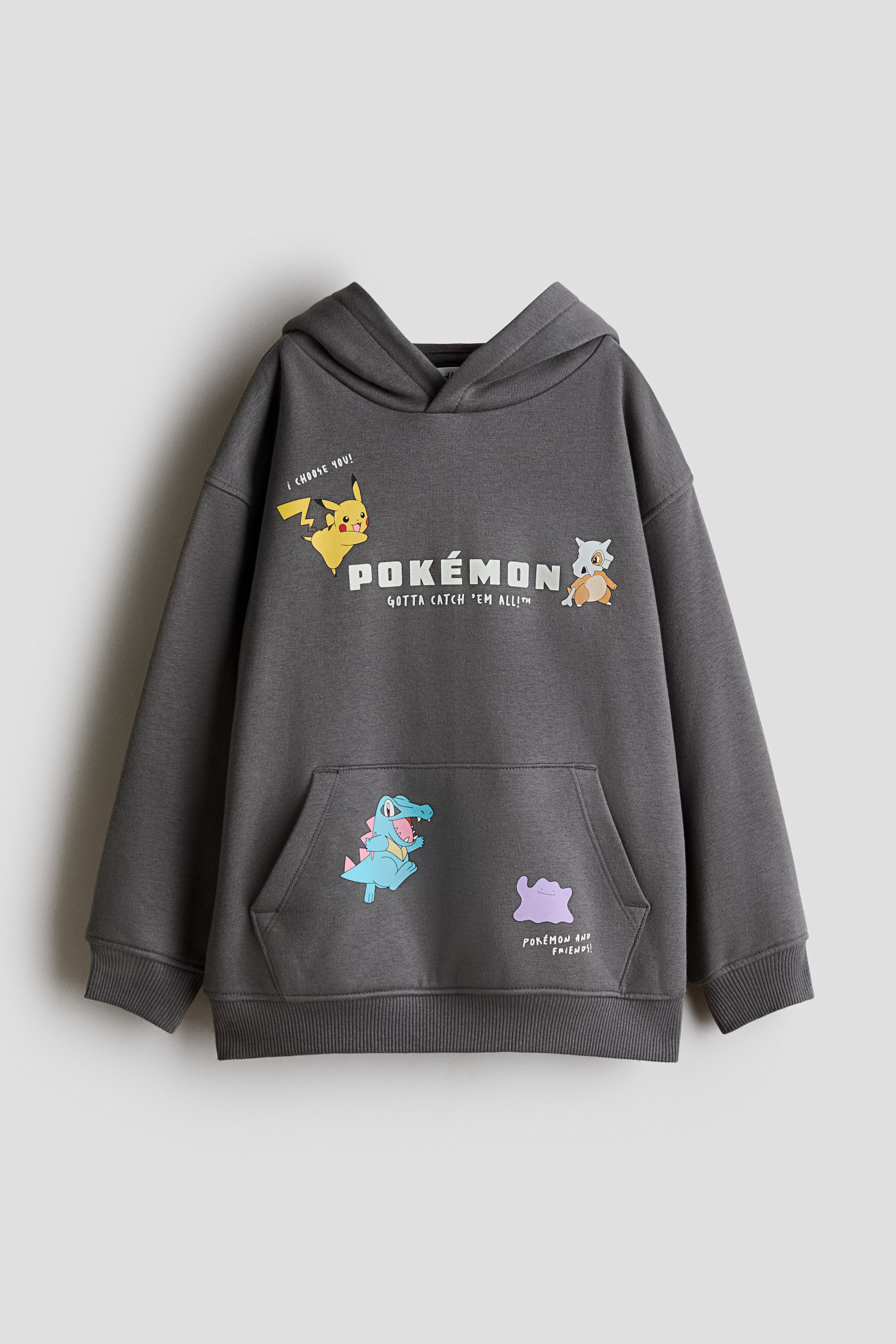 Agrandir l'image: Un hoodie gris foncé à coupe ample est présenté de face, doté d’une capuche doublée en jersey avec un col croisé sur le devant, de manches tombantes et d’une poche kangourou à l’avant. Le hoodie affiche des graphismes et du texte Pokémon imprimés sur la poitrine et la poche, et se termine par des poignets côtelés et un ourlet côtelé.
