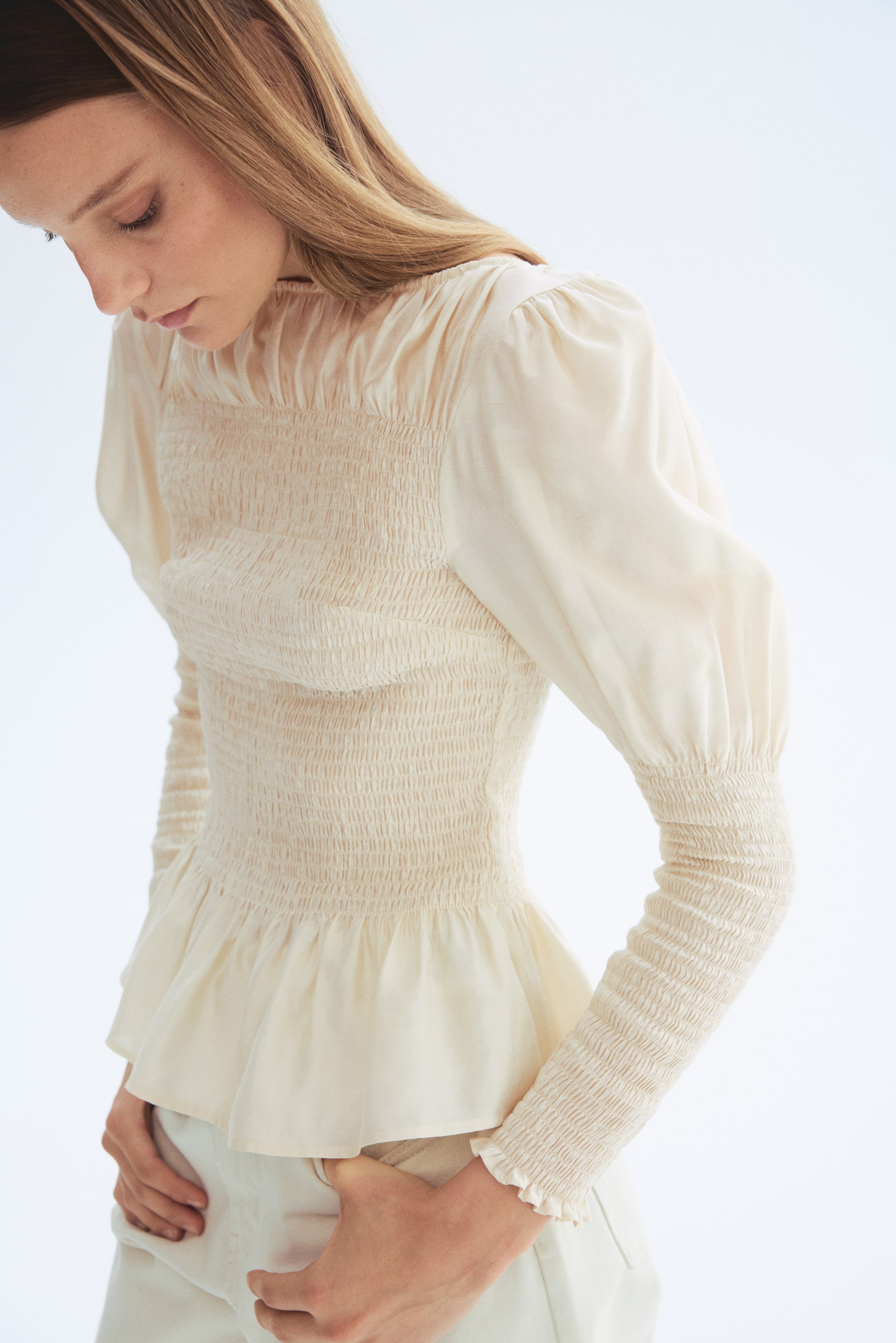 Ver imagem maior: Uma blusa creme é apresentada num ângulo ligeiramente lateral-frontal, destacando um extenso franzido ao longo do corpete e das mangas inferiores, criando uma silhueta ajustada. Apresenta uma gola alta franzida, mangas superiores bufantes e uma bainha peplum em evasé.