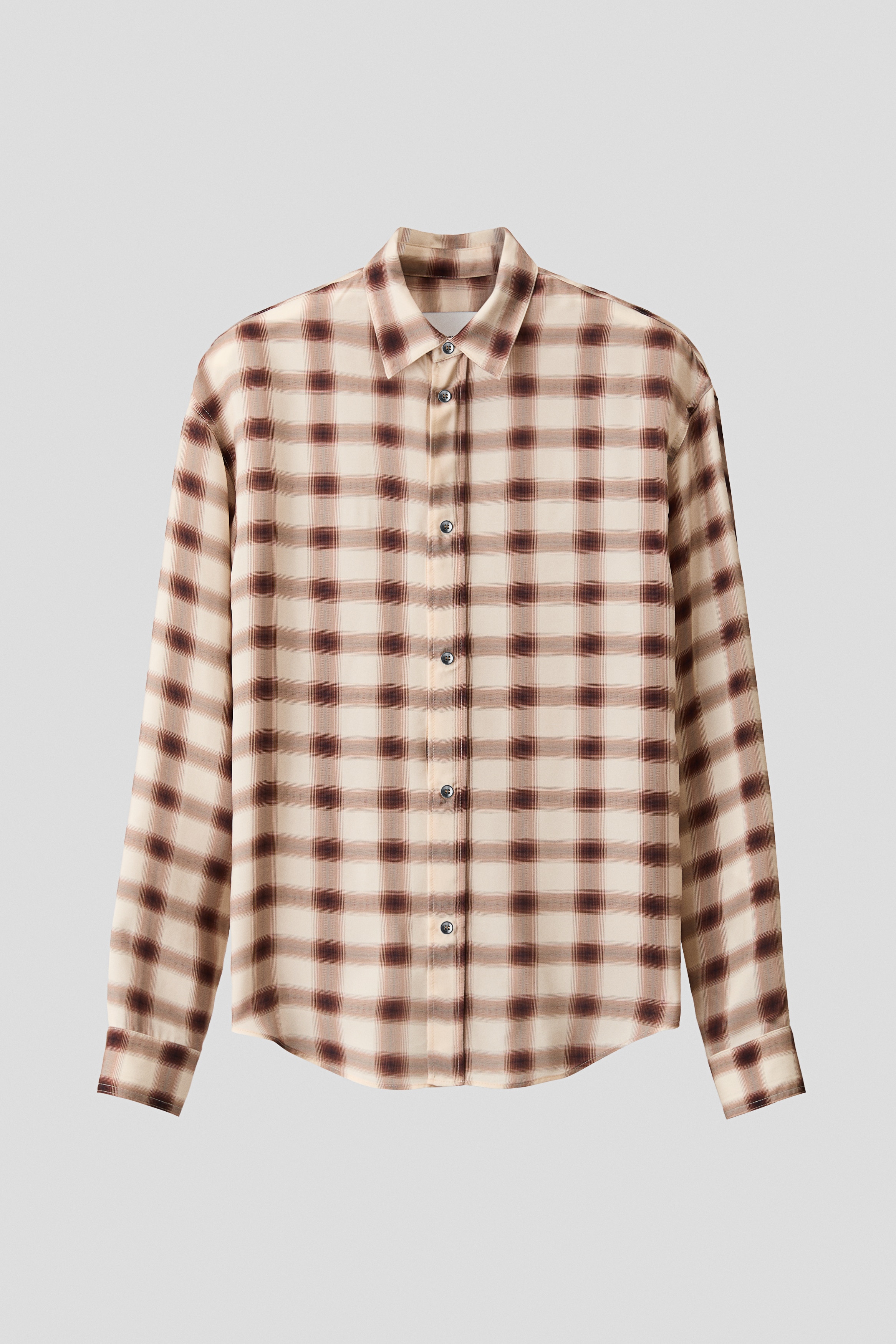 En långärmad skjorta i ett suddigt rutmönster i brunt och beige bärs av modellen, med ena sidan av fållen instoppad i mörkblå jeans. Skjortan har klassisk krage, knappslå och manschetter, och stylas med ett mörkt bälte och mörka skor.