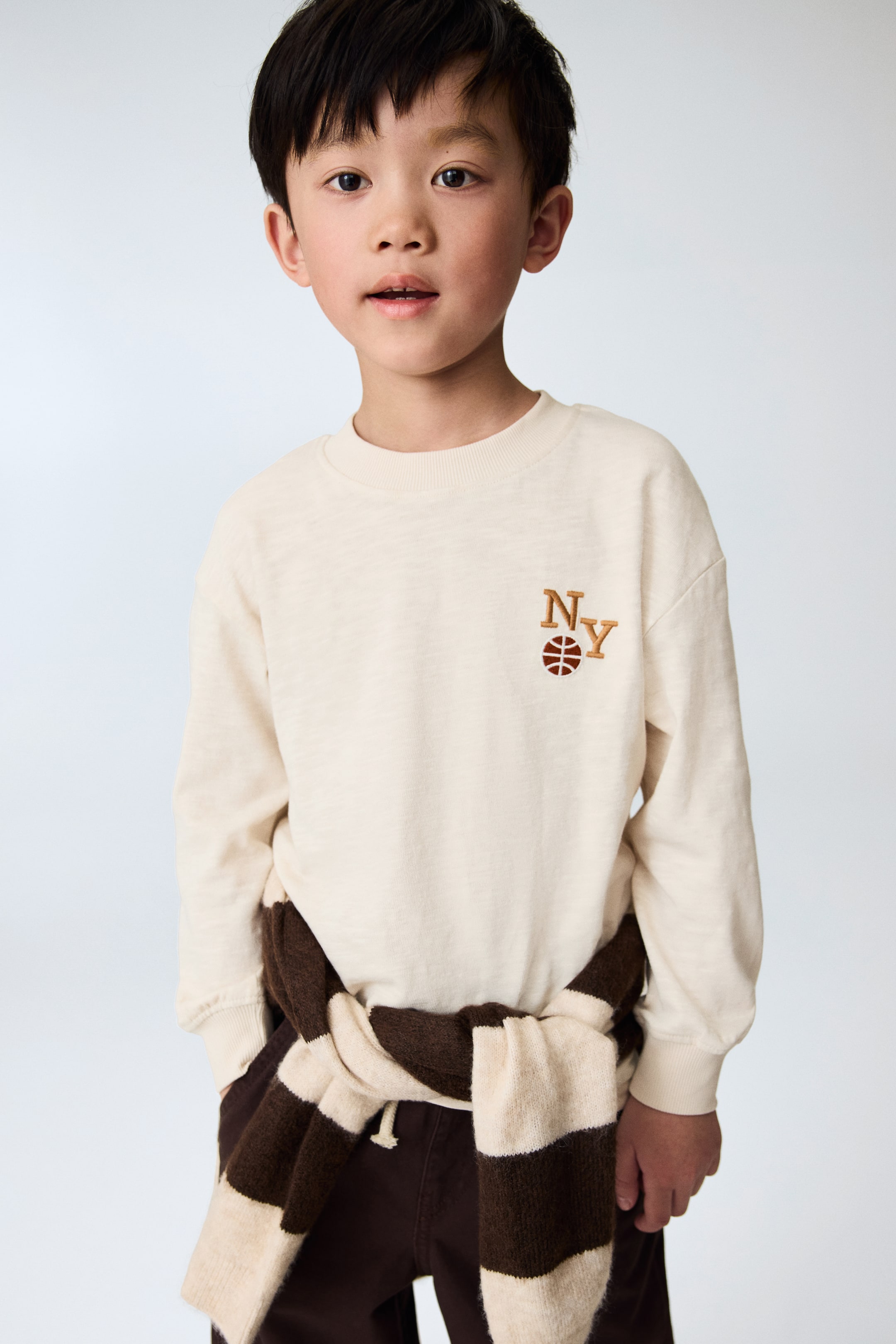 View larger image: An off-white long-sleeved top is worn, showcasing a ribbed crew neck, ribbed cuffs, and an embroidered "NY" lettering with a basketball icon in brown thread on the left chest. A brown and cream striped jumper is tied around the waist, styled over the top.