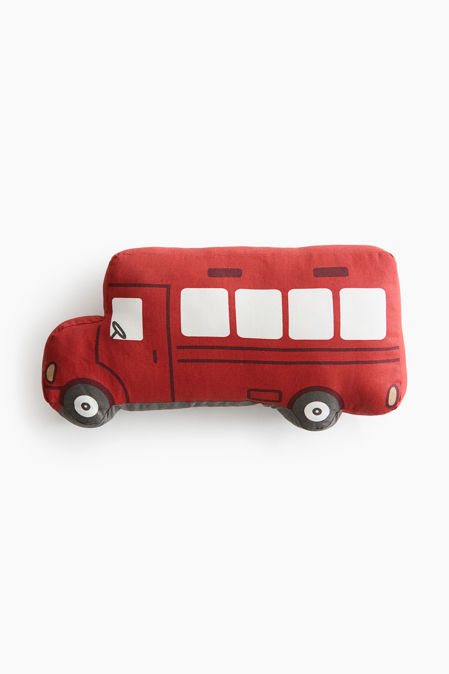 A red cotton canvas cushion shaped like a bus is shown from the front, featuring a playful vehicle design with printed details. The design includes four white passenger windows, a driver’s window with a steering wheel, and two contoured wheels along the bottom edge.