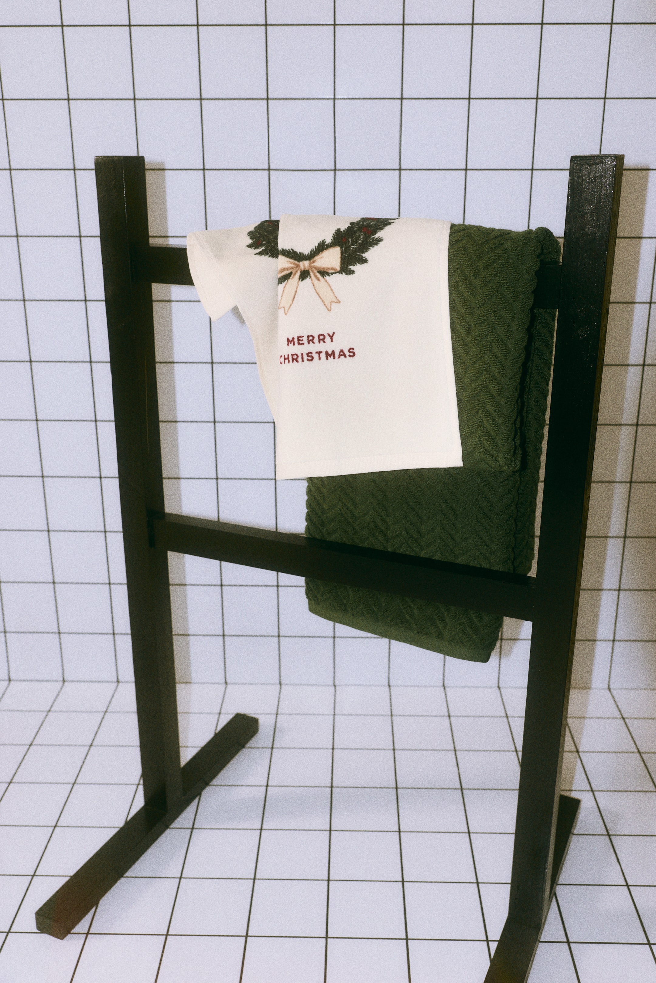 Grotere afbeelding bekijken: Een paar handdoeken hangt over een zwart rek, met daaronder een getextureerde donkergroene handdoek en daarboven een gebroken witte badstof handdoek van katoen. De gebroken witte handdoek toont een bedrukt ontwerp op de voorkant, met een groene dennenkrans met een lichtgekleurde strik en de tekst 'MERRY CHRISTMAS' in rode letters.