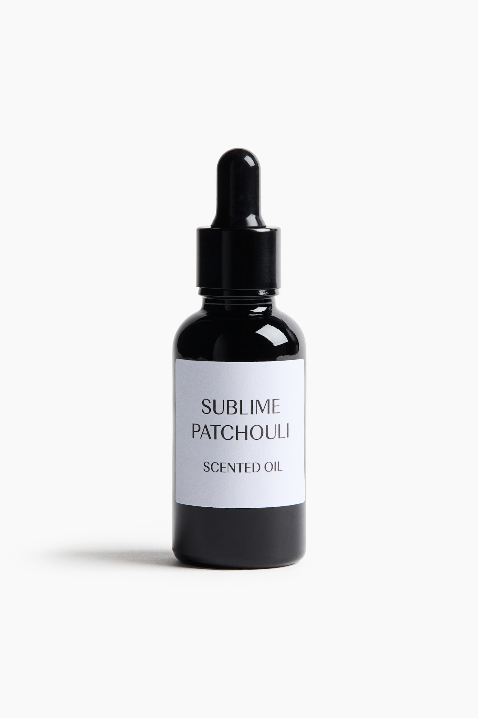 En sort glasflaske med pipette står oprejst og vender fronten fremad, med en minimalistisk hvid rektangulær etiket med teksten "SUBLIME PATCHOULI" og "SCENTED OIL" trykt i sort. Flasken er udstyret med en matchende sort pipettehætte. - 1