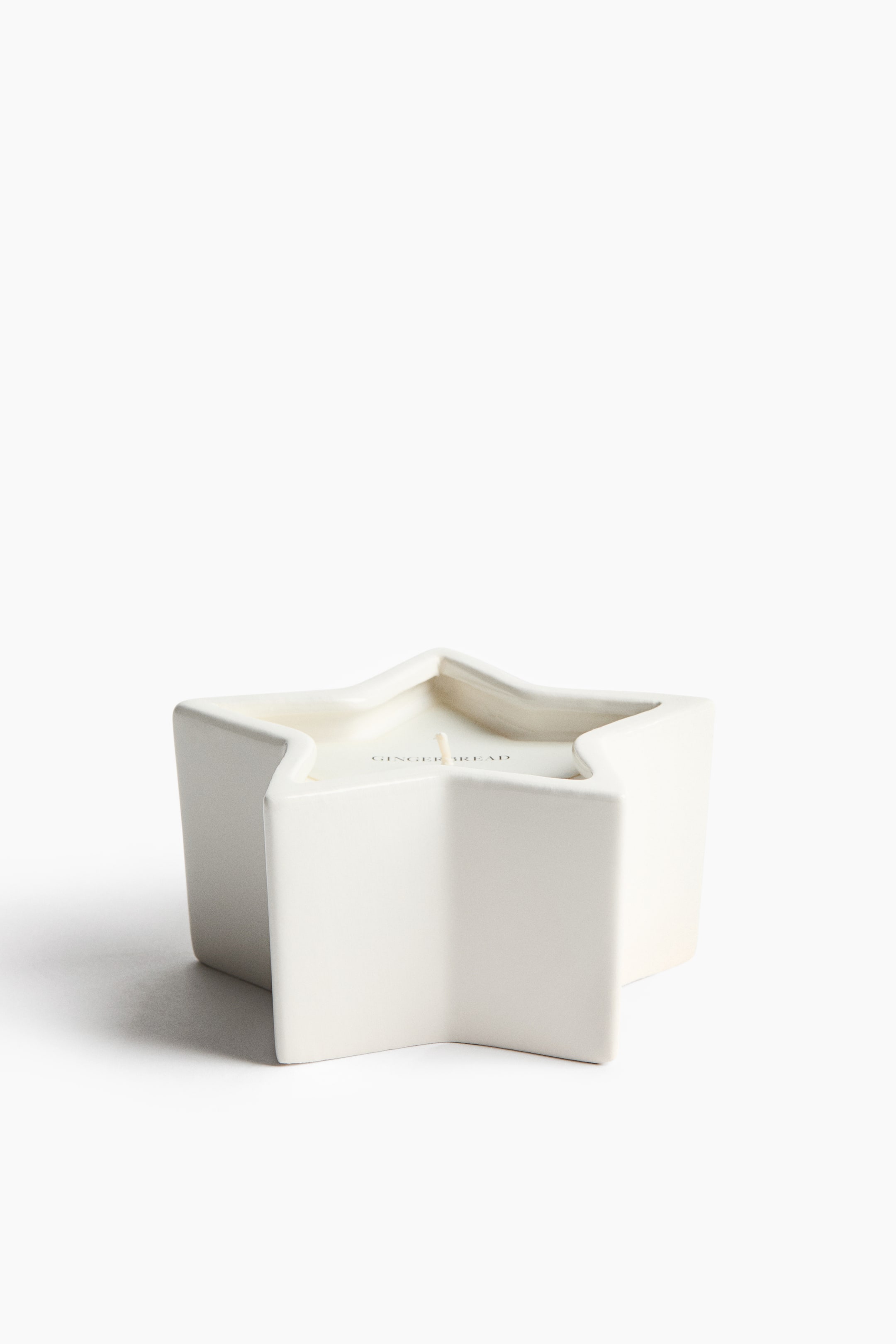 View larger image: A star-shaped candle features a white ceramic container with a five-pointed star design, shown from the front at a slightly elevated angle. The container holds white wax with a single central wick and the word "GINGERBREAD" printed on the wax surface.