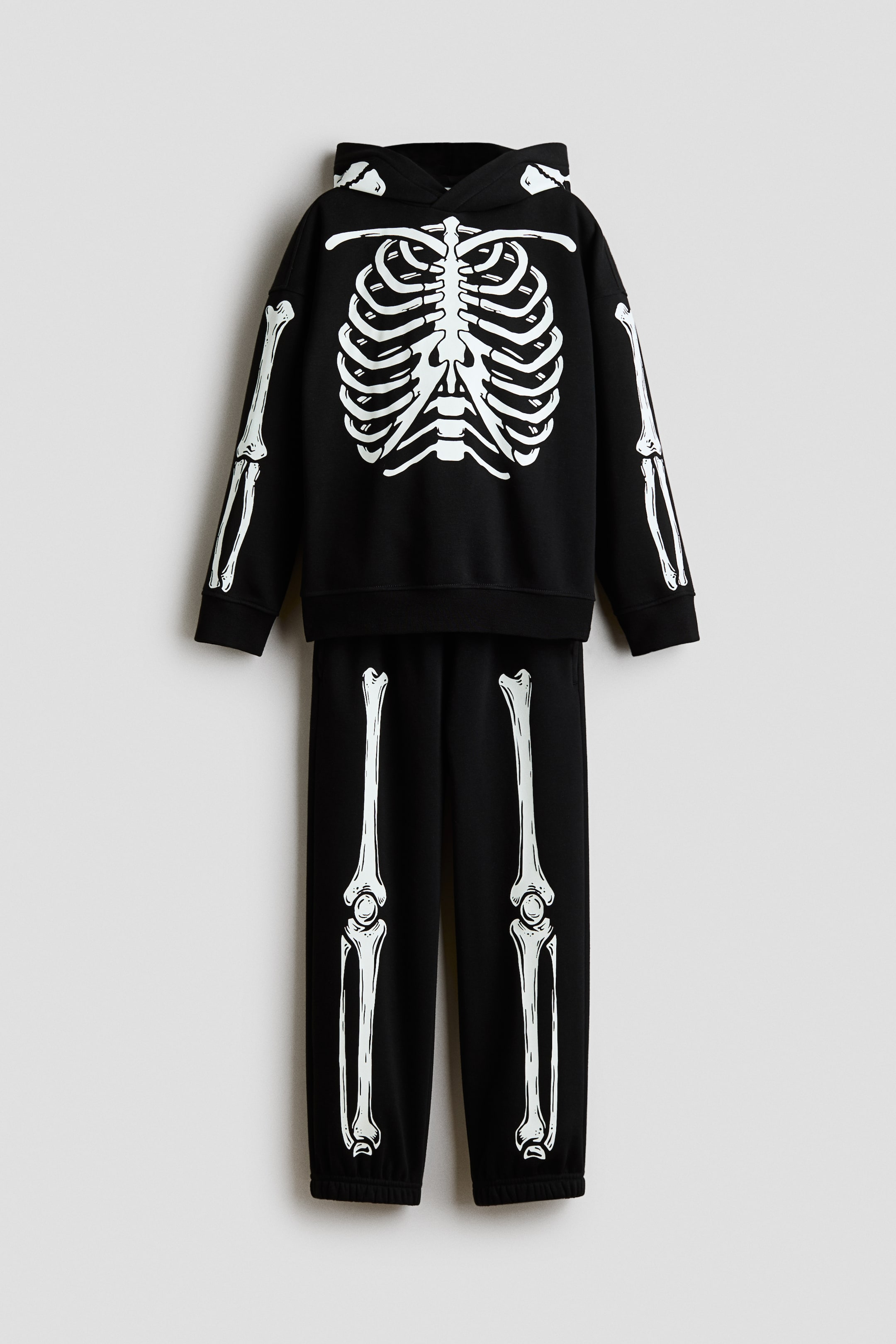 View larger image: This front-facing two-piece set features a black hoodie and matching sweatpants with a white skeleton print. The hoodie displays a ribcage design on the chest and arm bones on the sleeves, while the sweatpants feature leg bone prints running down the front of each leg.