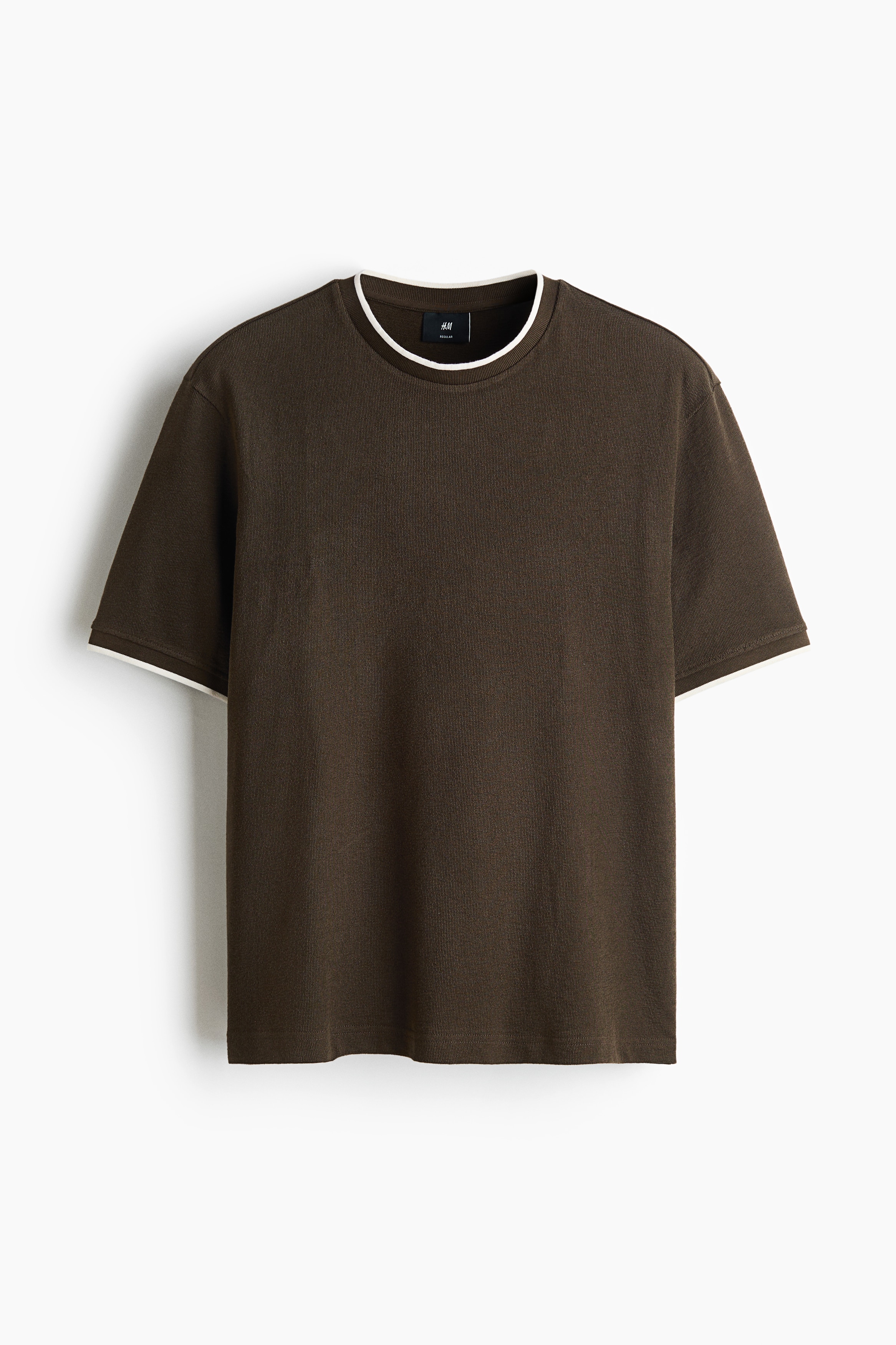 The model is wearing a brown heavyweight cotton jersey T-shirt with contrasting cream-coloured trims at the neckline and sleeve cuffs. The regular-fit T-shirt features a straight hem and is paired with off-white trousers.