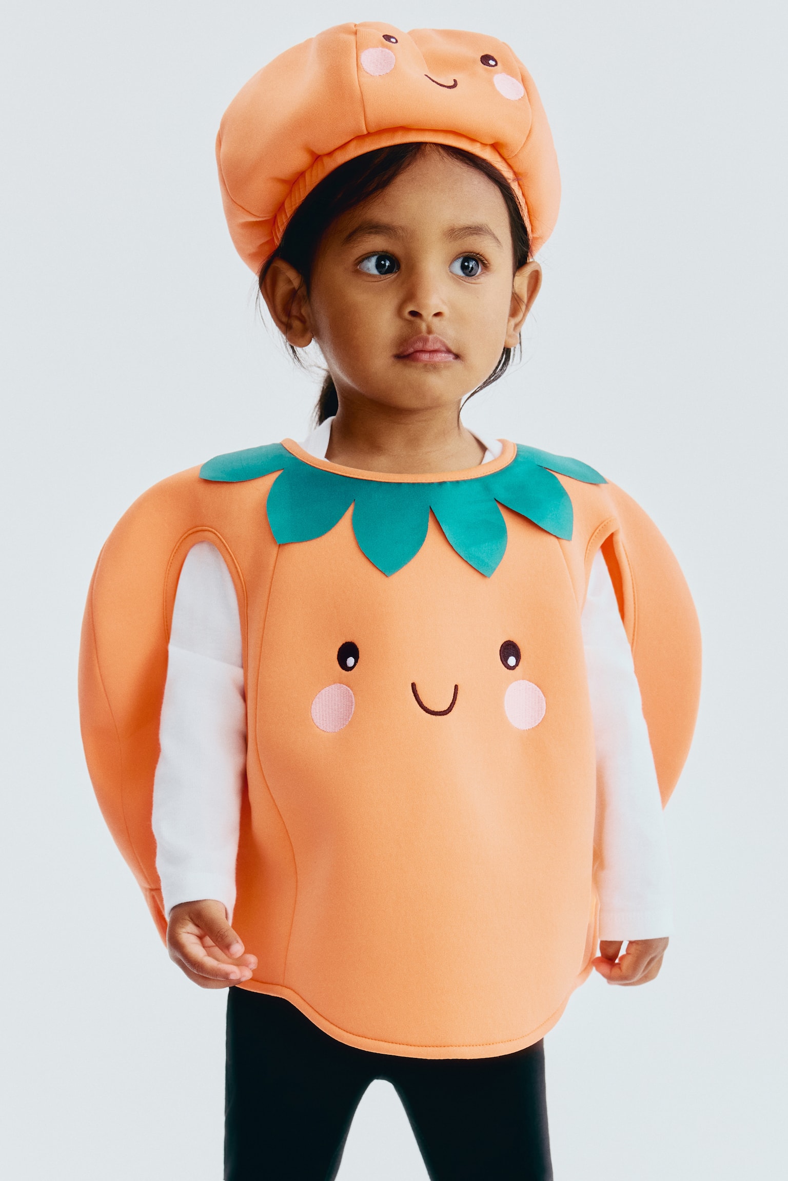 Un costume de déguisement élégant, en forme de citrouille orange, est posé à plat et présente un col en forme de feuille verte autour de l'ouverture du cou. Il arbore des yeux noirs brodés, une bouche souriante et des cercles de fard à joues roses sur le devant, avec deux ouvertures ovales pour les bras découpées sur les côtés. - 1