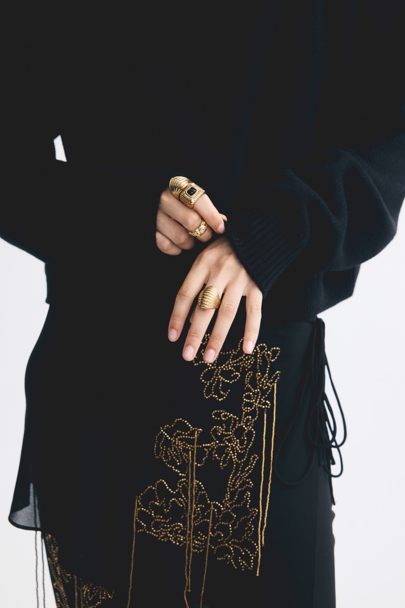 Rings in a variety of designs, including three in metal and one in plastic, are being worn on both hands by the model. The model is lightly holding the ribbed sleeve of a black knit sweater, highlighting a chunky gold-toned ring with a black square insert and a textured band on one hand, and a ribbed dome-shaped gold ring on the other. - 1
