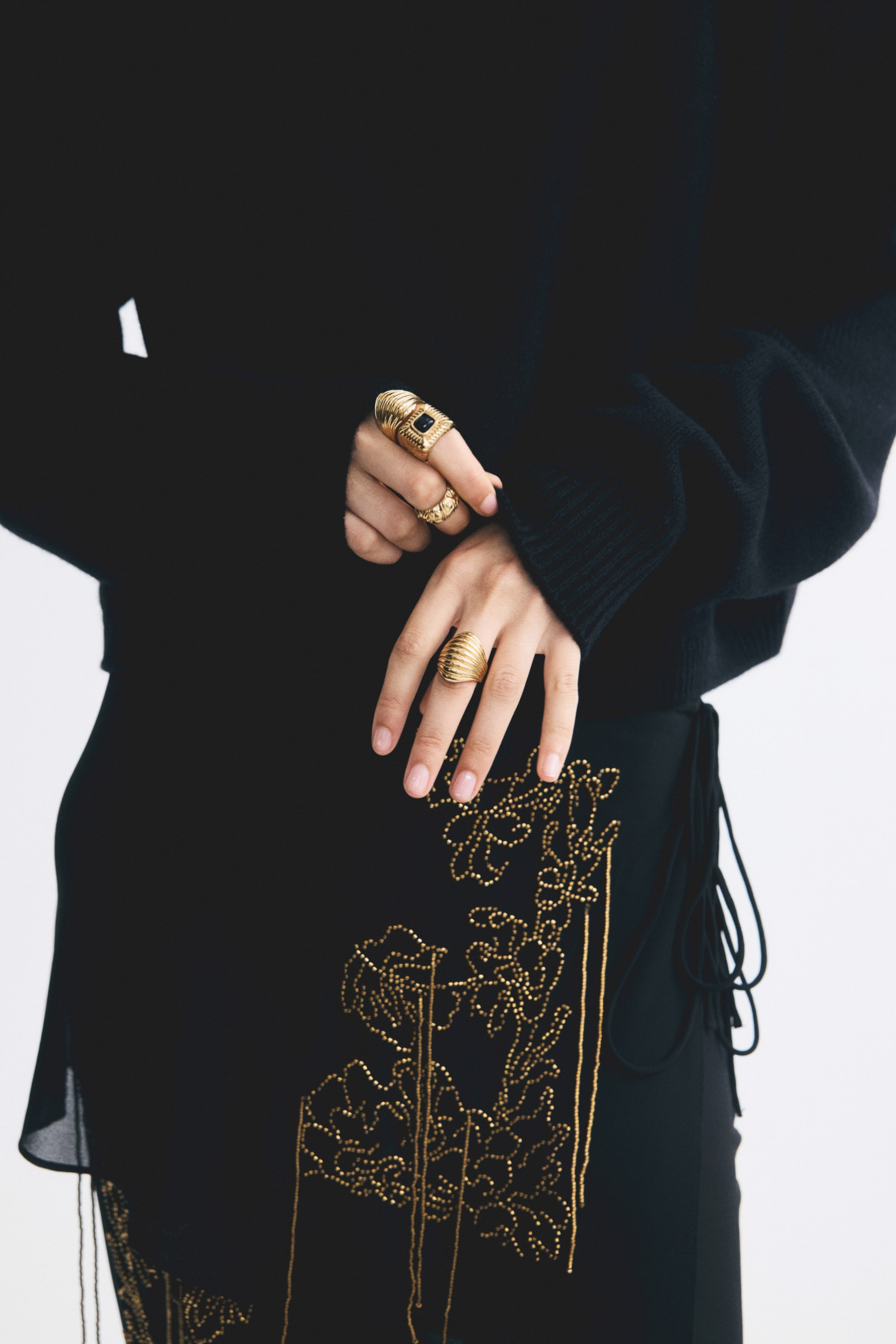 View larger image: Rings in various designs, including three in metal and one in plastic, are being worn on both hands by the model. The model is lightly holding the ribbed sleeve of a black knit sweater, highlighting a chunky gold-toned ring with a black square insert and a textured band on one hand, and a ribbed dome-shaped gold ring on the other.
