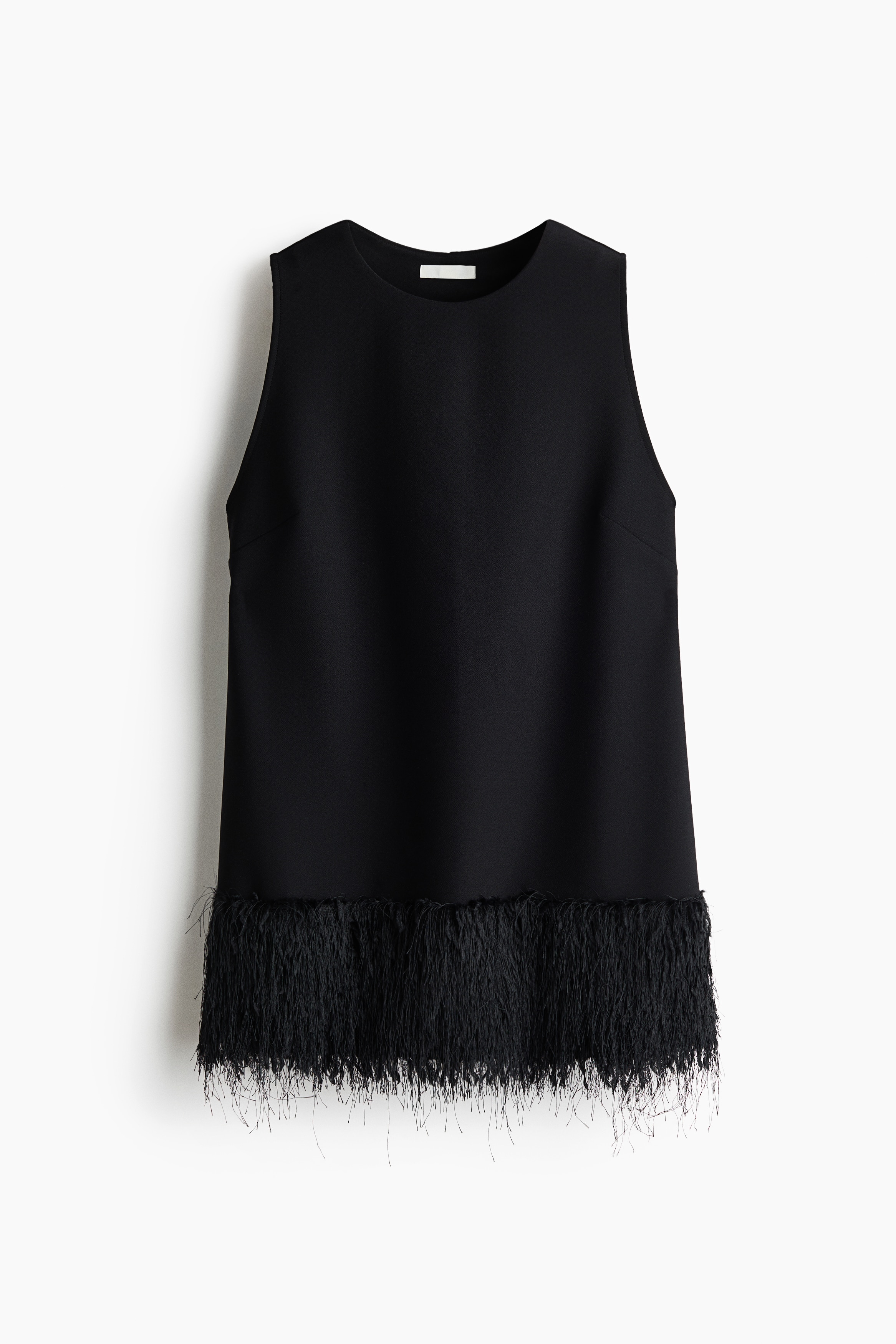 A sleeveless black top featuring a loose fit, a round neckline and textured fringe at the hem is worn by the model crouching in profile. The top is styled with matching black trousers featuring similar fringe trim at the cuffs and black pointed-toe heeled shoes.