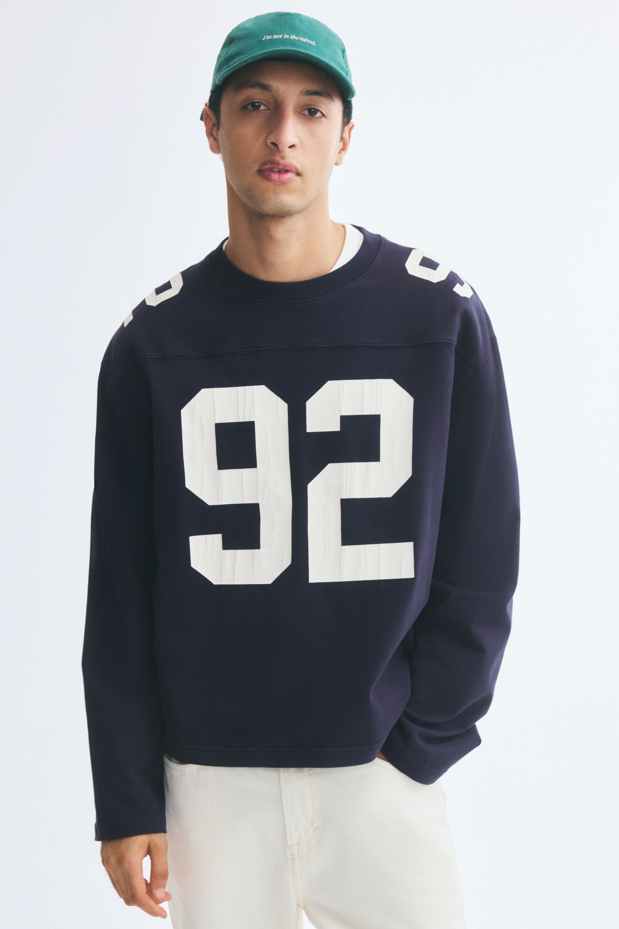 View larger image: A dark blue long-sleeve tee in a relaxed fit with a front yoke is being worn by the model. The tee features a large white number "92" printed on the chest, matching white prints on the shoulders, and a ribbed round neckline layered over a white collar.