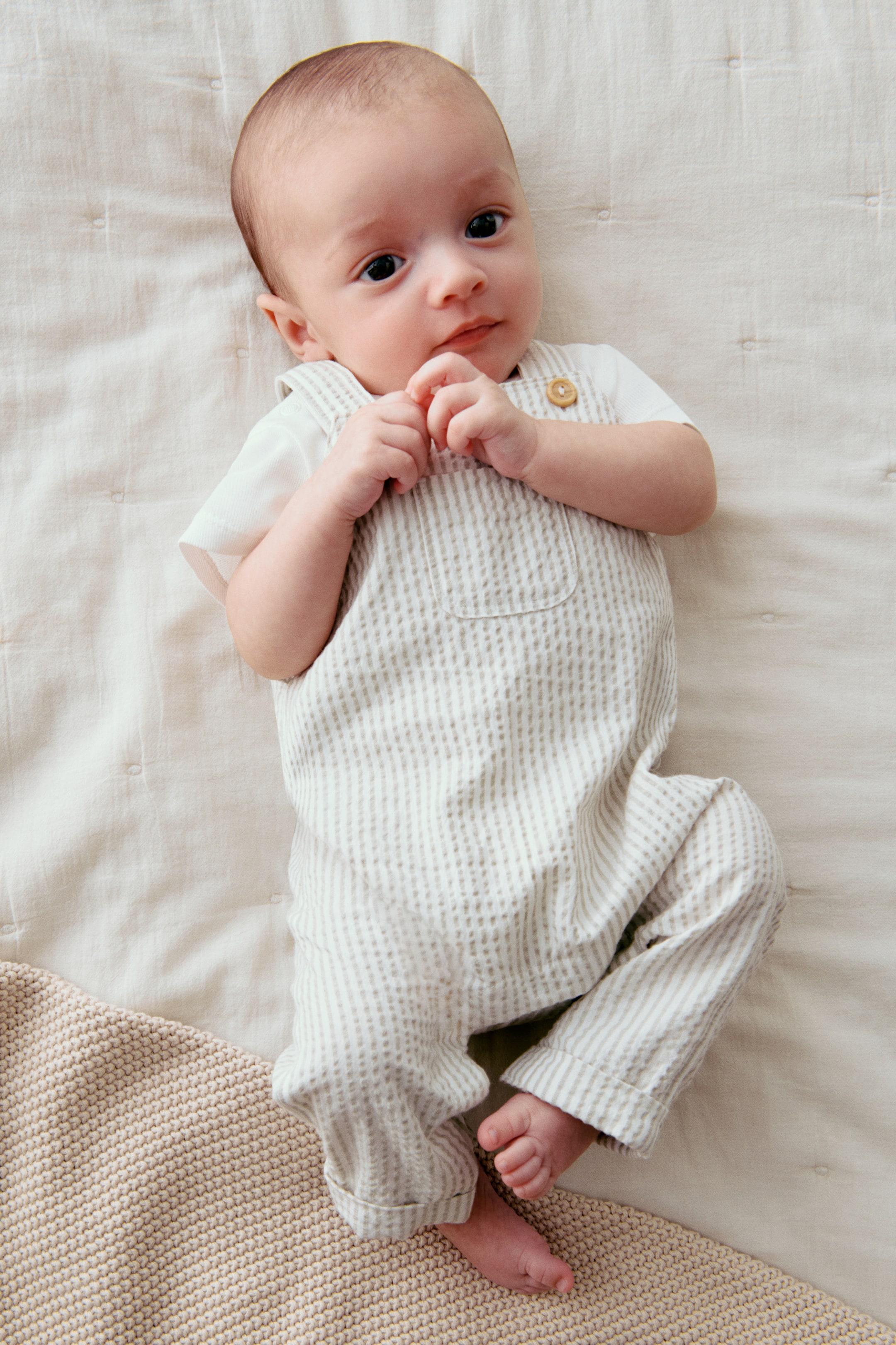 View larger image: A light-colored, vertically striped baby overall romper is being presented from the FRONT. It is featuring a full-length design with a bib, shoulder straps, a round wooden button on the right strap, and a small patch pocket on the bib, layered over a white short-sleeved t-shirt.