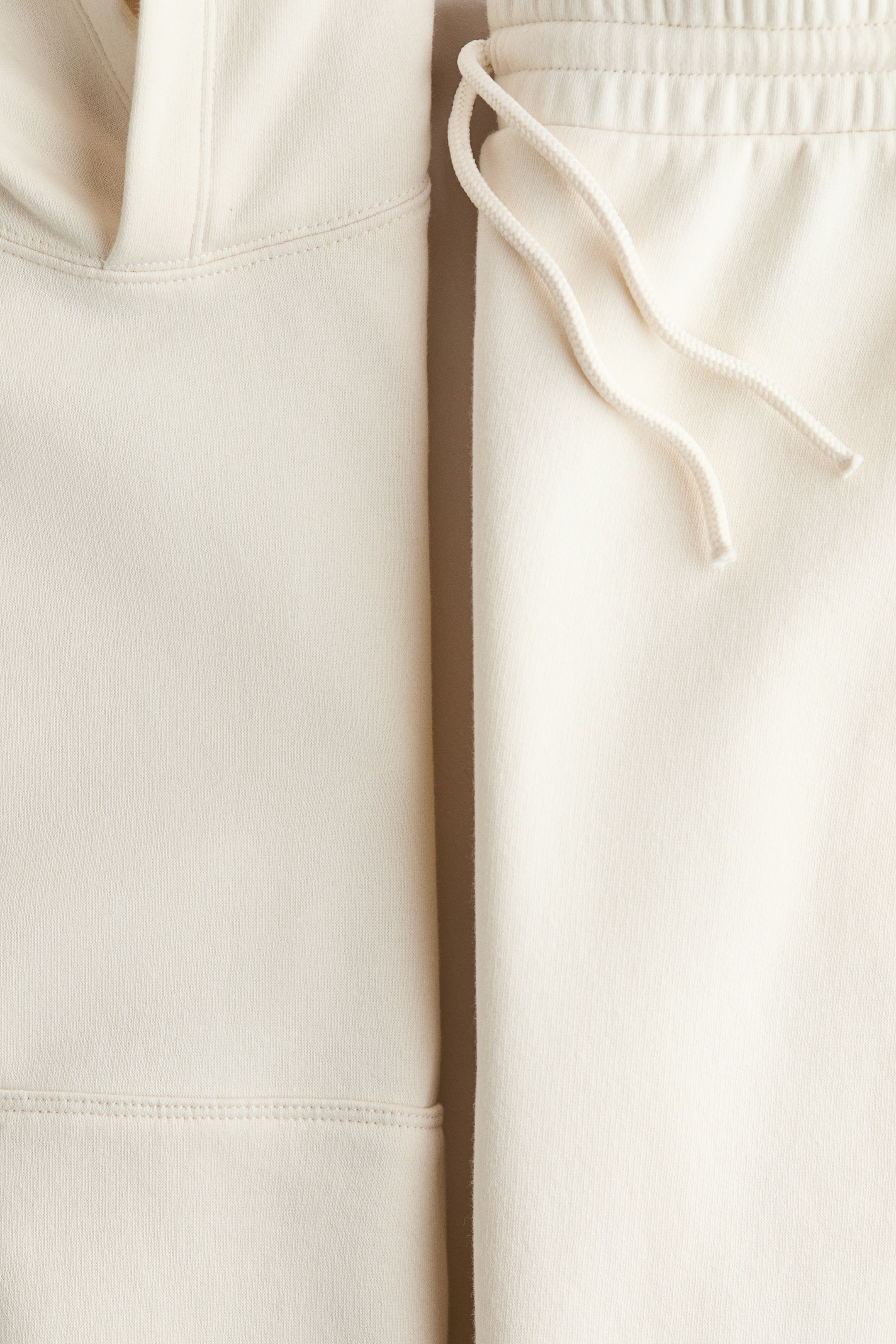 View larger image: Two cream-colored, soft jersey garments are presenting a cohesive look, with a hooded top featuring a visible opening and stitched seam on the left, and a matching bottom showcasing an elasticated waistband and drawstring on the right.