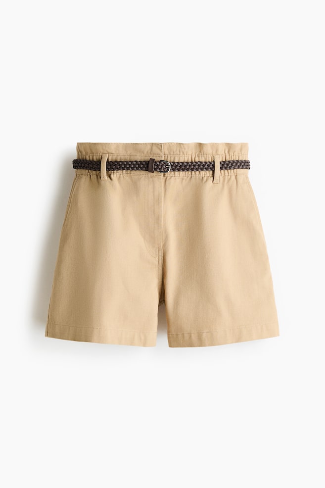 Tan shorts are featuring a gathered waistband with belt loops and visible front pockets. A dark brown braided belt with a silver-toned buckle is threaded through the loops. - 1