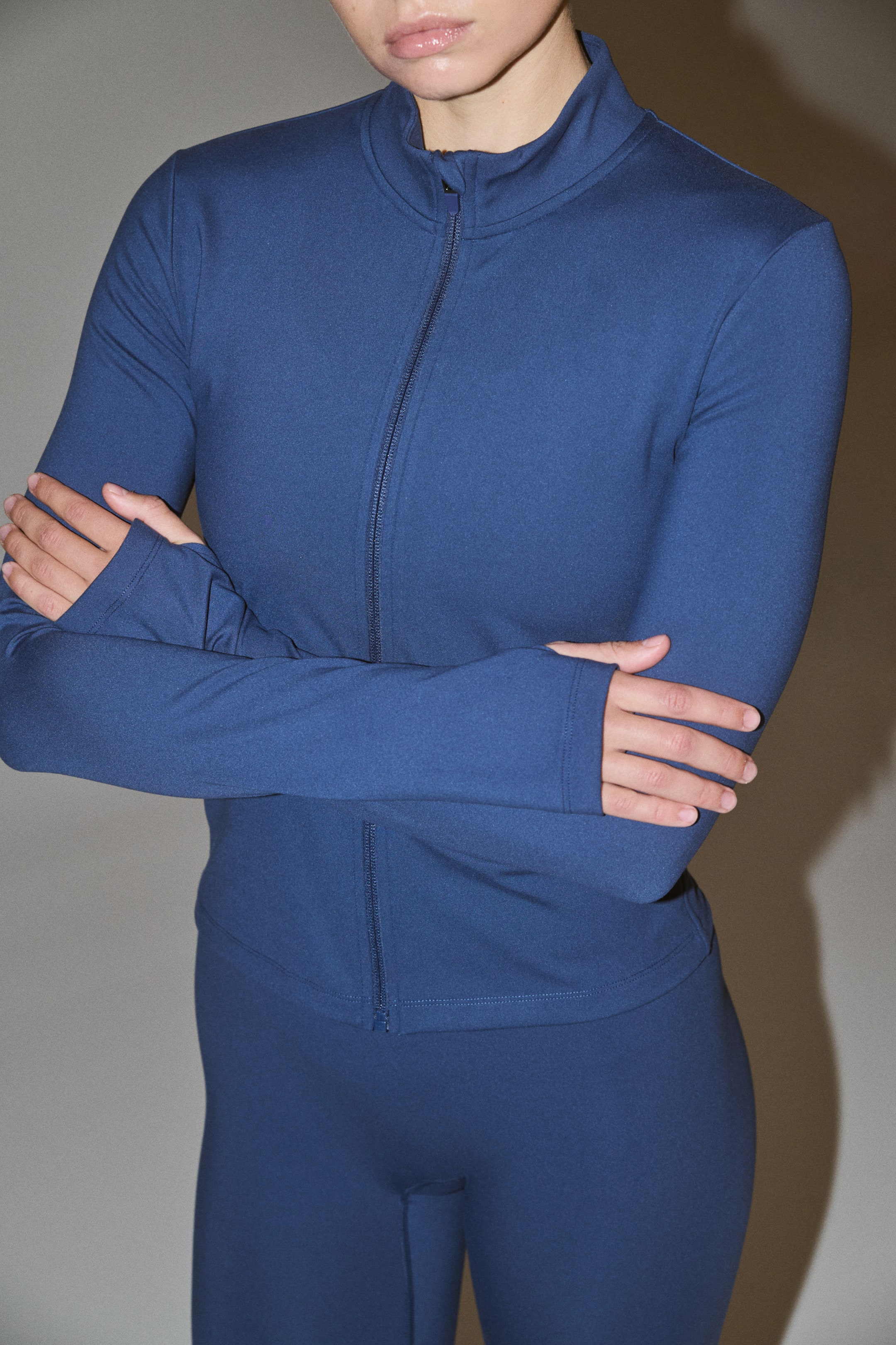 Ampliar la imagen: Un top deportivo azul oscuro con cierre completo y leggings a juego conforman un conjunto de ropa deportiva entallado. El top presenta un cuello tipo mock, mangas largas con aberturas para el pulgar en los puños y un detalle de costura lateral visible.