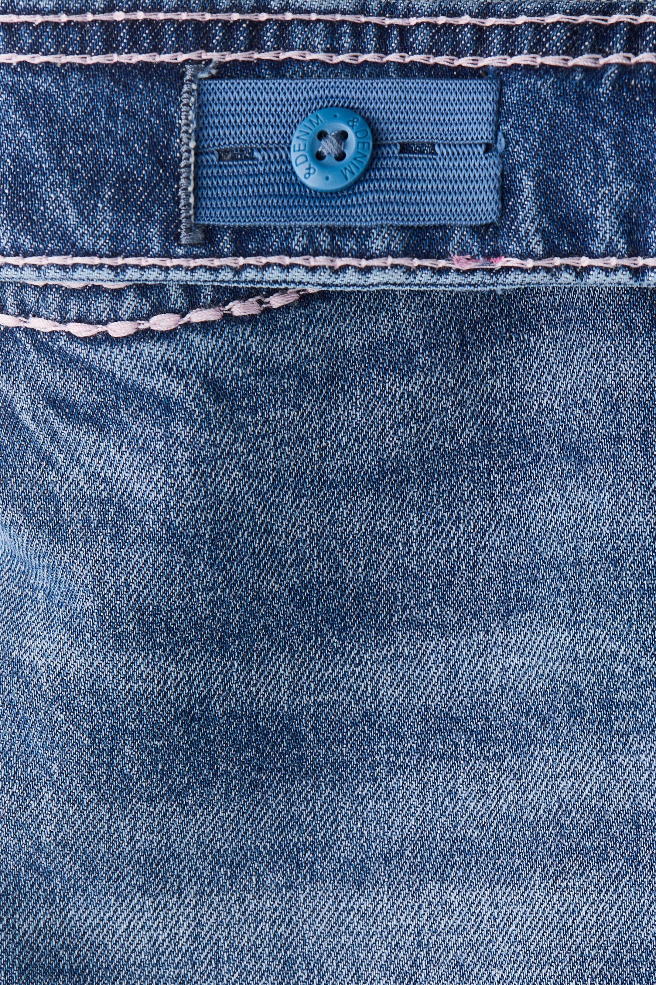 View larger image: A pair of blue denim jeans is showcasing an adjustable elastic waistband on the FRONT, with light pink stitching detailing a horizontal seam. A blue elastic tab with buttonholes is visible, and a blue button, embossed with 'DENIM', is fastened through one of the buttonholes.