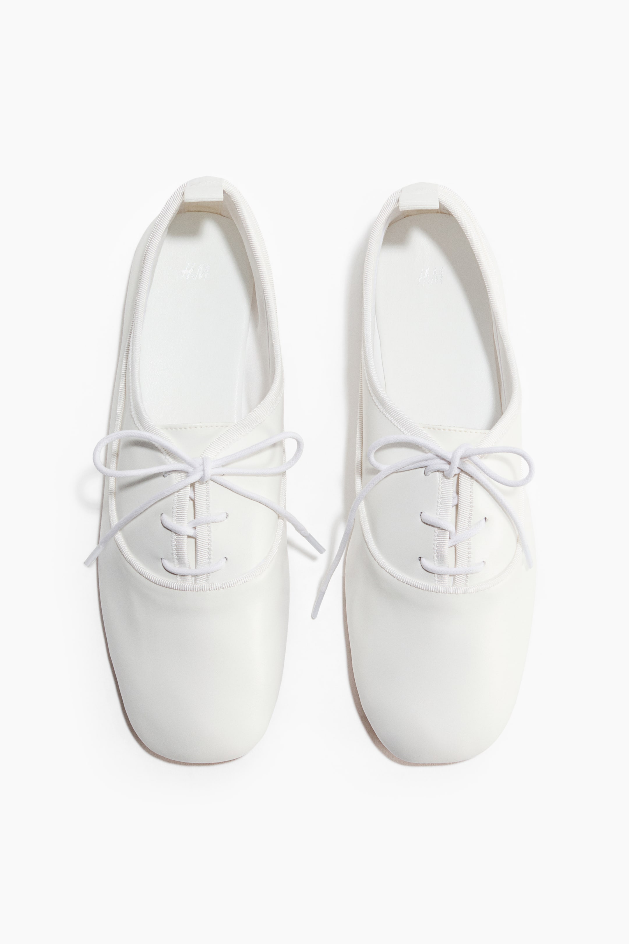 Ver imagen más grande: Un par de zapatos planos blancos muestra un diseño minimalista, con ambos zapatos orientados hacia el frente. Cada zapato presenta una parte superior lisa y mate con cuatro pares de ojales blancos, a través de los cuales se han pasado cordones blancos finos, atados en un lazo.