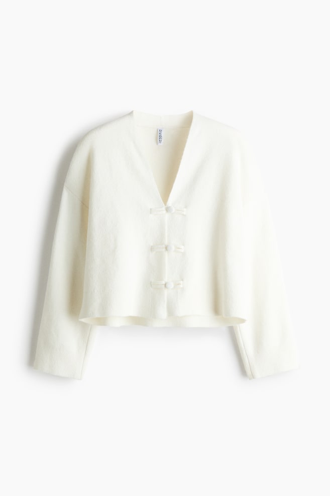 A cream-coloured knit cardigan is hanging in a front view, showcasing a deep V-neckline, dropped shoulders, and relaxed wide sleeves. The slightly cropped cardigan fastens down the centre with three fabric-loop frog closures and matching buttons. - 1