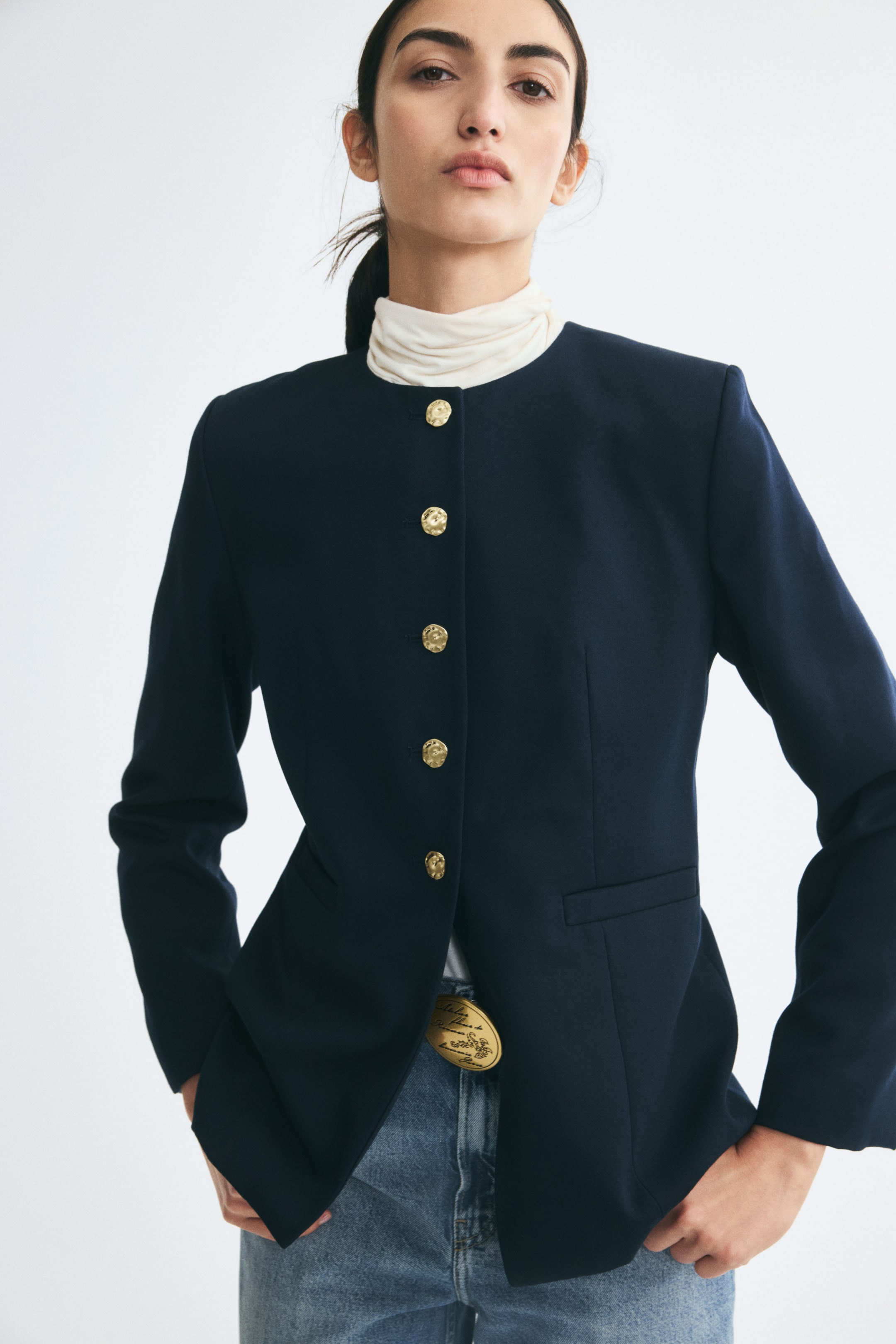View larger image: The model is wearing a navy blue, single-breasted twill blazer featuring a round neckline and five irregular-shaped gold buttons. The structured blazer is layered over a cream turtleneck and paired with light-wash blue jeans.