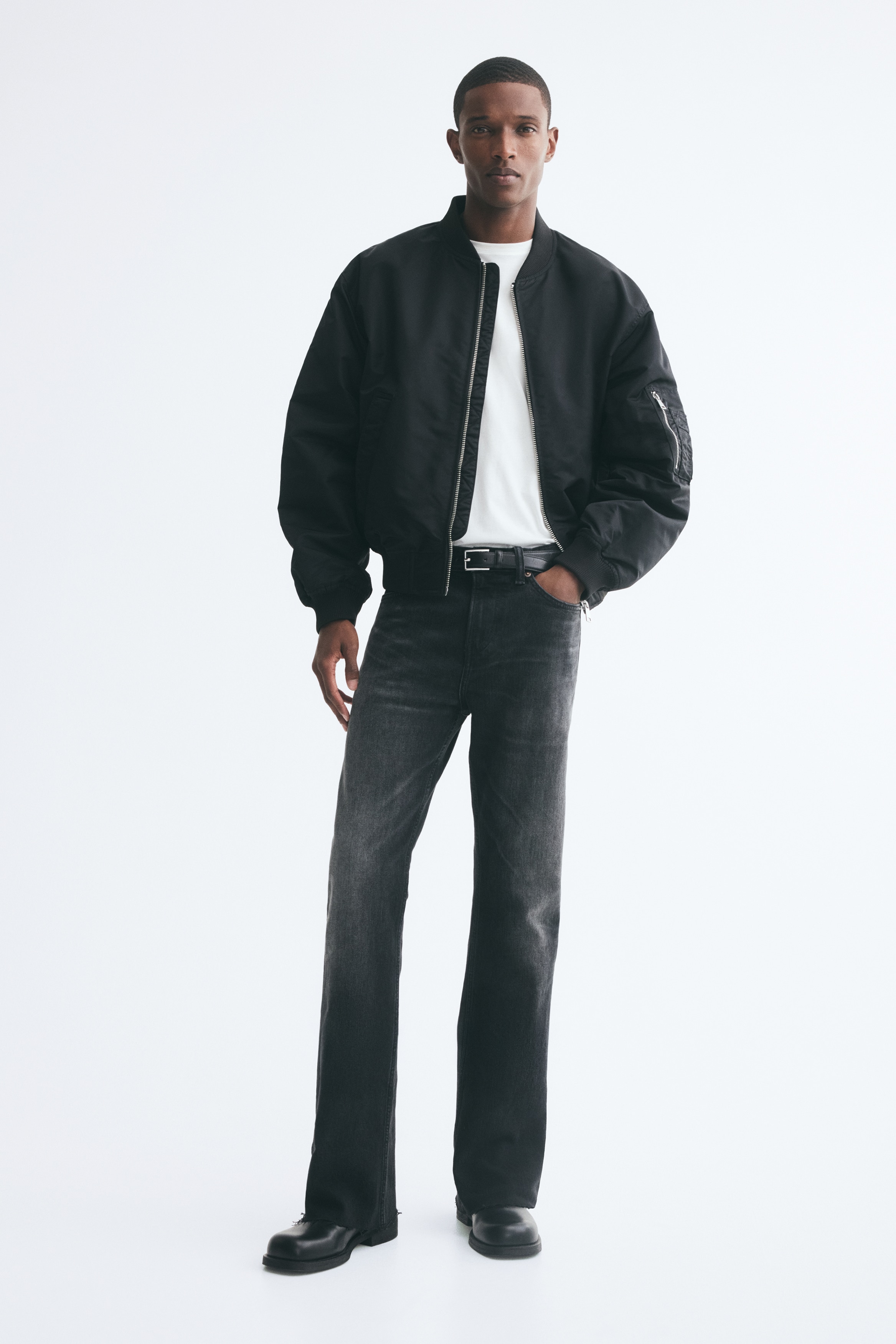 Ett par svartblekta jeans sedda framifrån, med rak benvidd, diskret whiskering vid höfterna och blekning ned över låren. Jeansen har en klassisk femficksdesign, bälteshällor, silverfärgad knappstängning och råa, fransiga fållar.