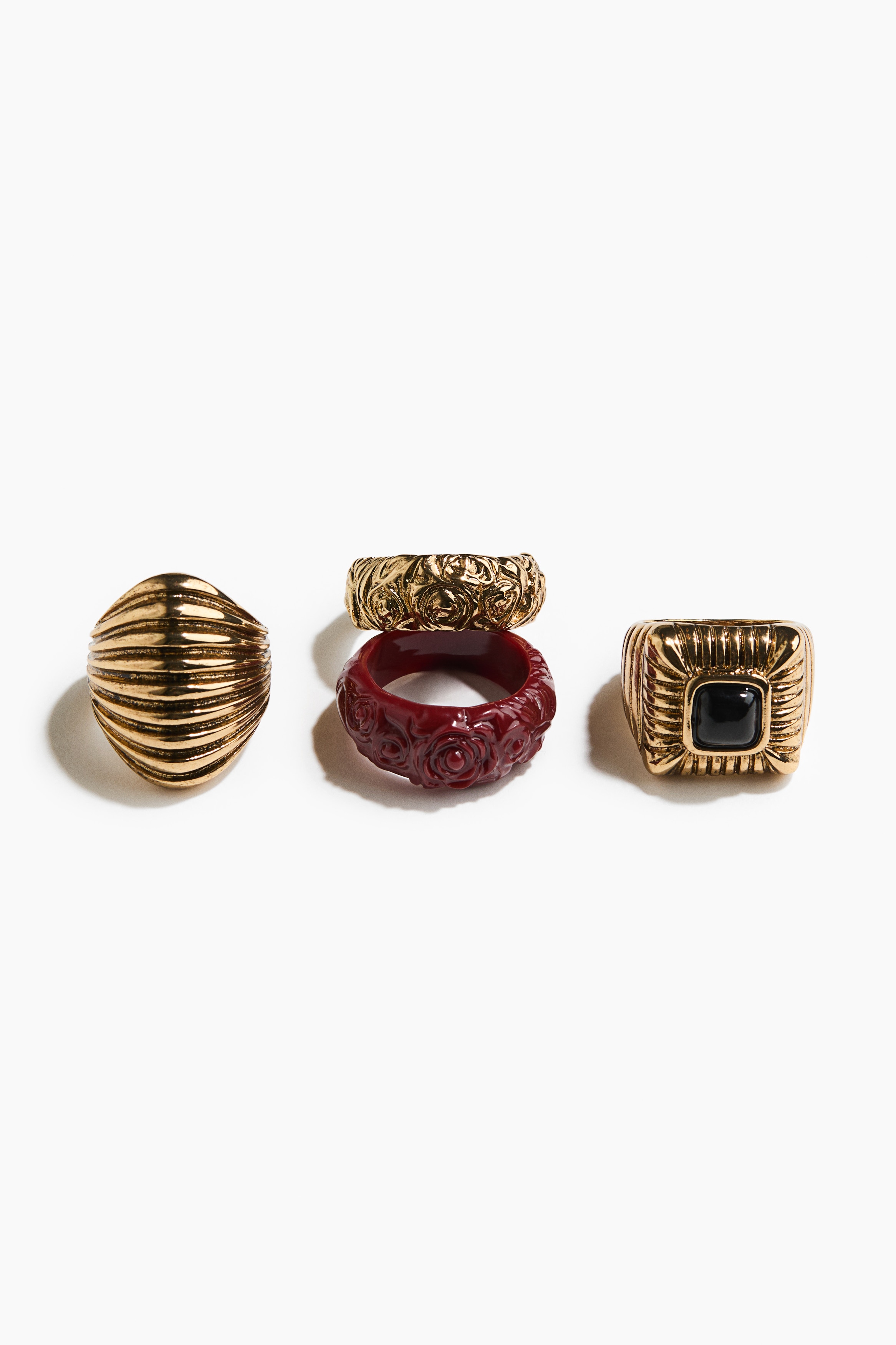 Rings in various designs, including three in metal and one in plastic, are being worn on both hands by the model. The model is lightly holding the ribbed sleeve of a black knit sweater, highlighting a chunky gold-toned ring with a black square insert and a textured band on one hand, and a ribbed dome-shaped gold ring on the other.