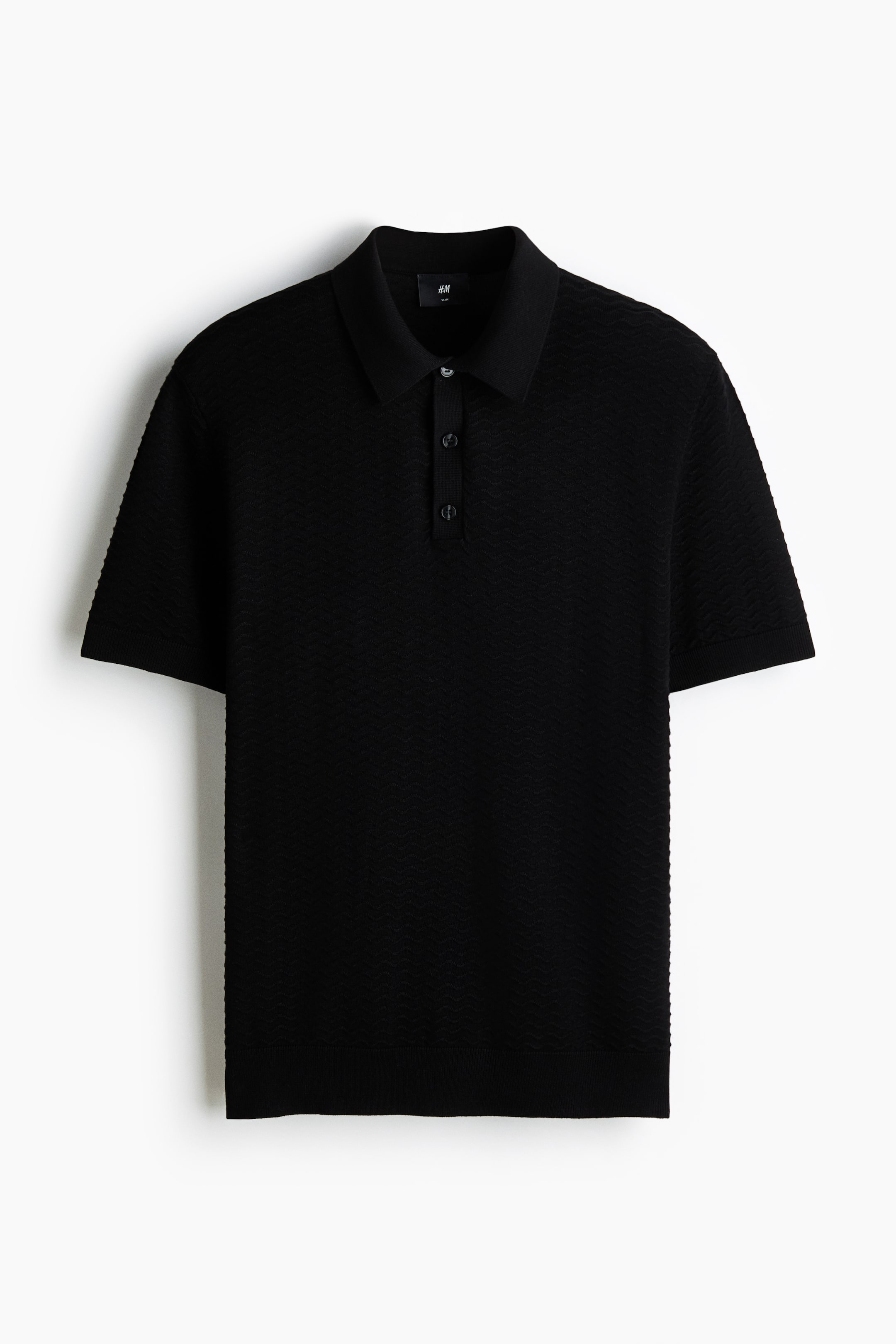 Ampliar la imagen: Una camiseta polo negra muestra su parte delantera, con una sutil textura de tejido ondulado en toda su superficie. Está diseñada con un cuello acanalado, una tapeta de tres botones, mangas cortas con puños acanalados y un dobladillo acanalado.