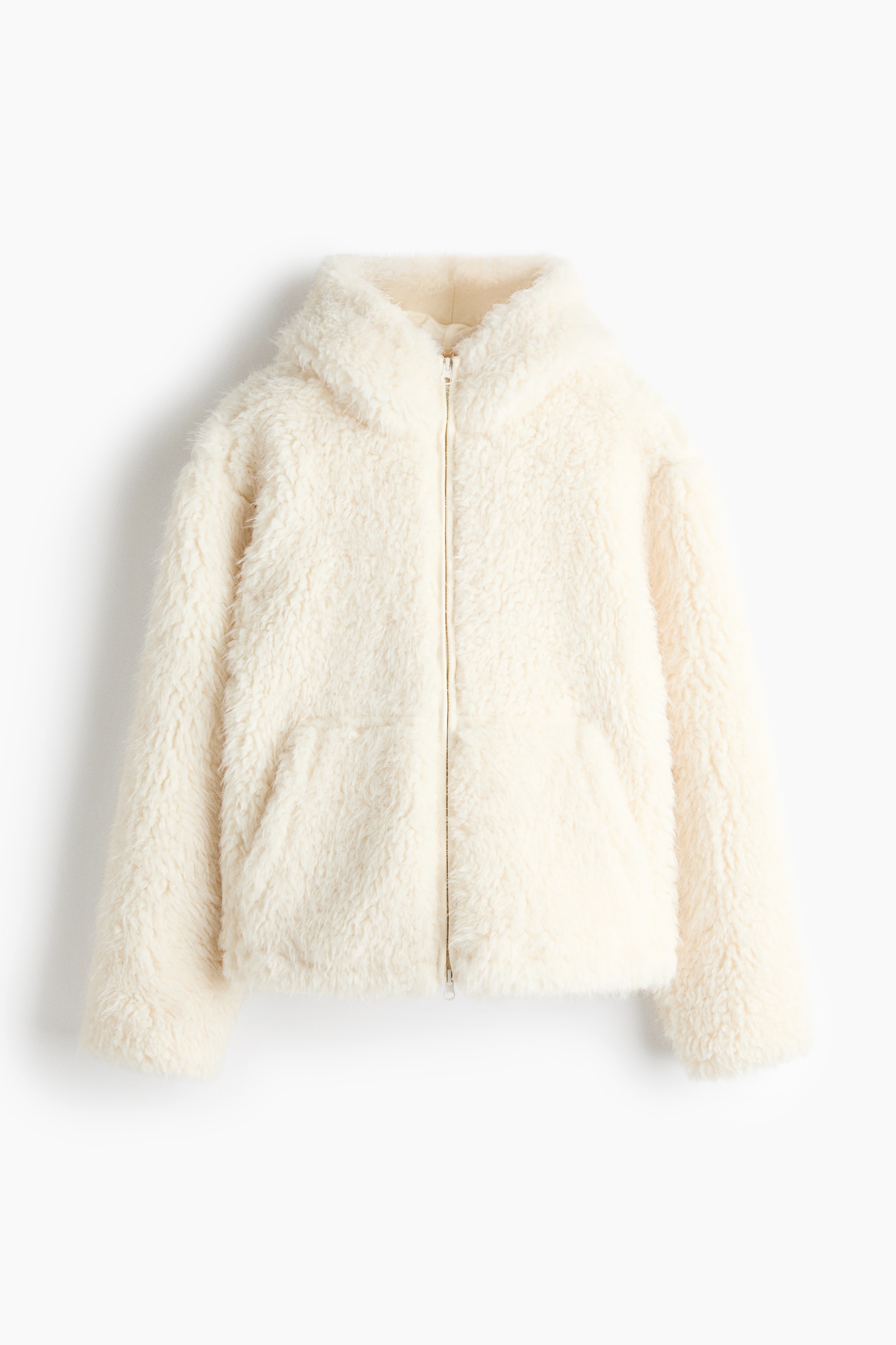 A cream-colored, textured faux-shearling puffer jacket is being worn by the model with their hands placed inside the front pockets. The jacket features a high-collar hood and a full-length front zipper closure.