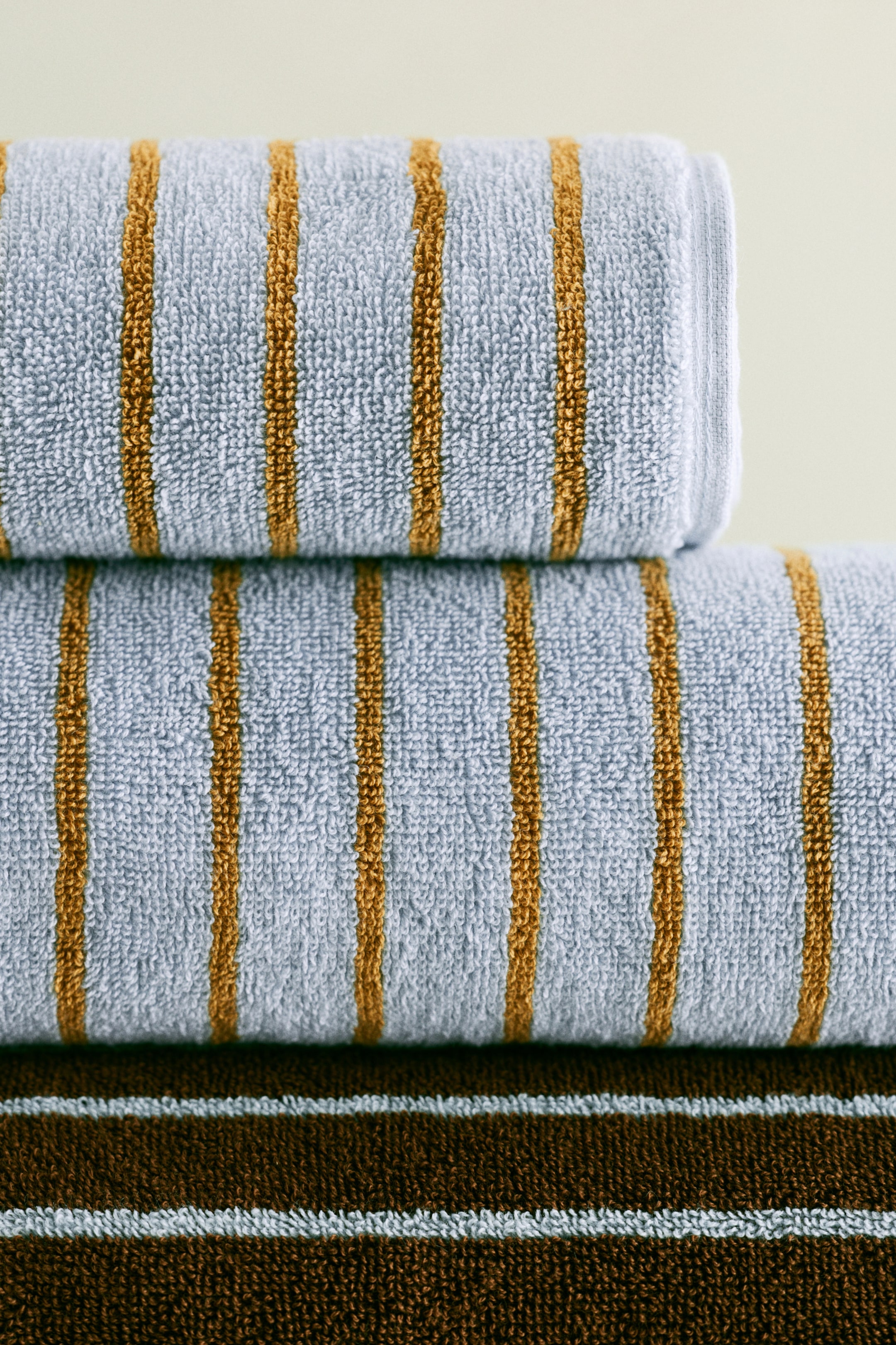 View larger image: A stack of terry cloth towels is showcased, with the top two light blue towels adorned with golden-brown vertical stripes. The bottom dark brown towel features light blue horizontal stripes.