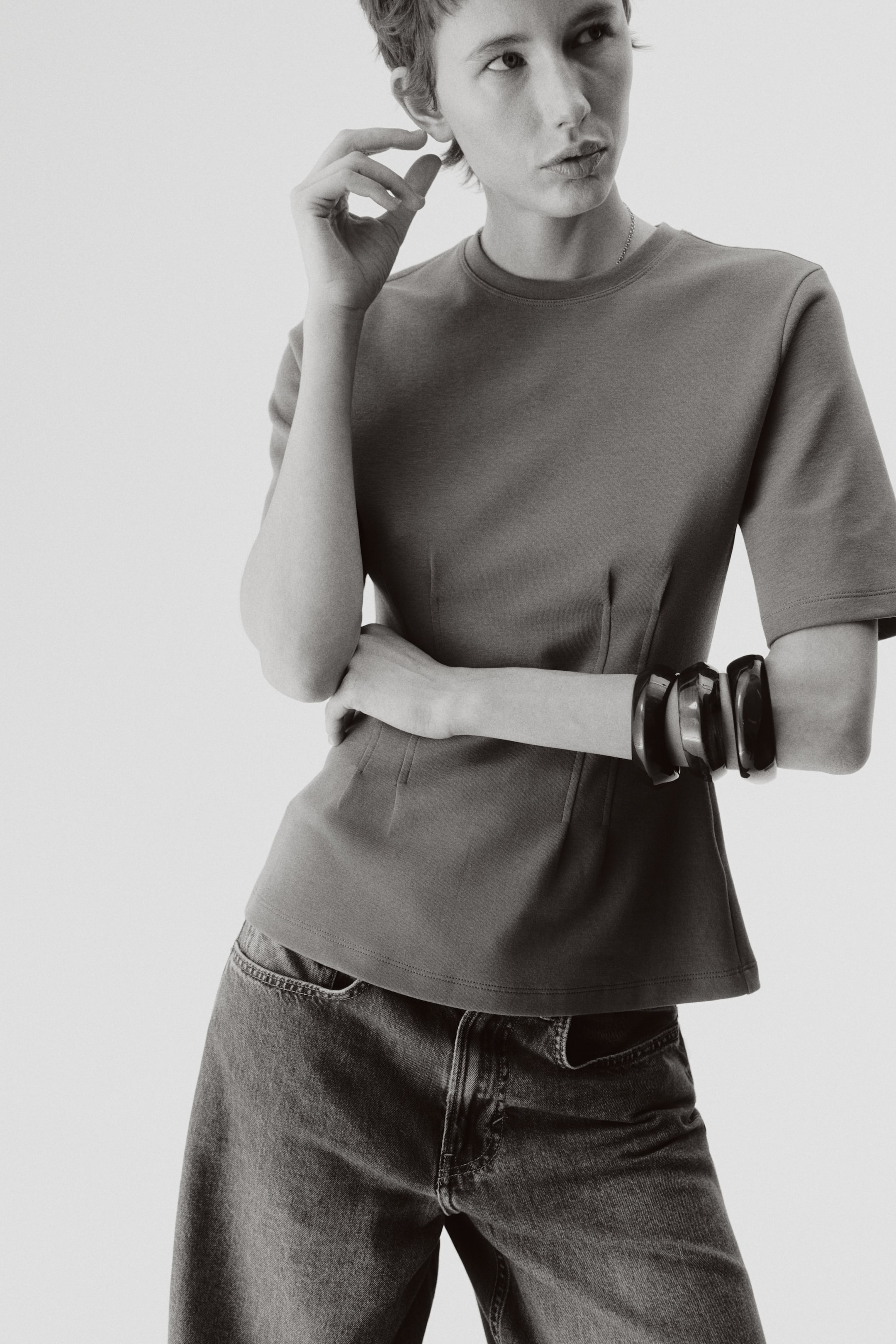 View larger image: A dark-toned, crew-neck top features short sleeves and a cinched waist with visible vertical seams, flaring slightly at the hem. Black bangles adorn the right sleeve.