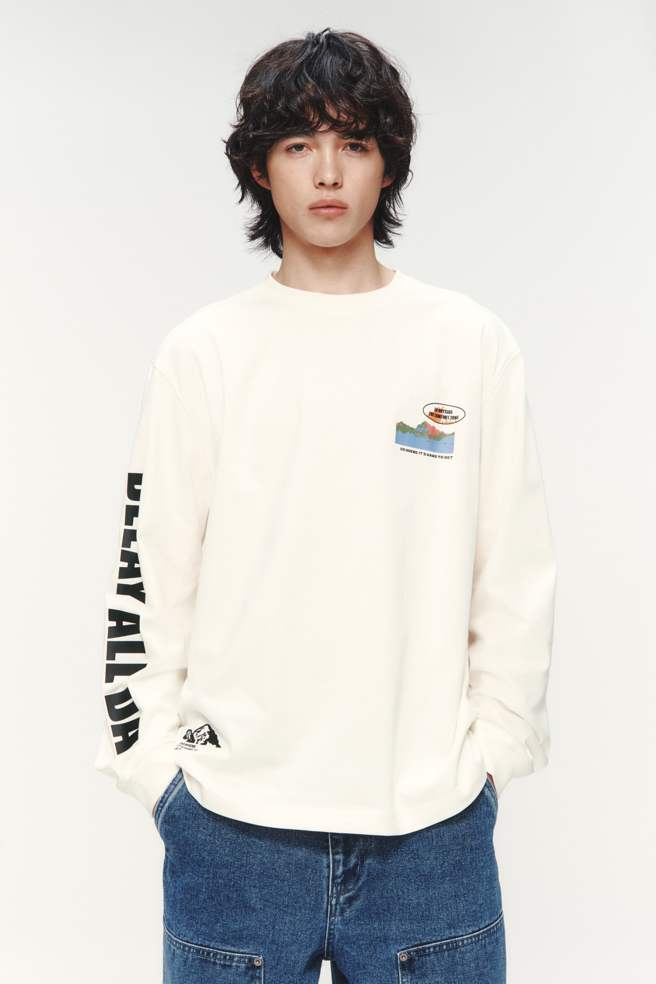 拡大画像を表示: モデルは、オフホワイトのルーズフィットで長袖のトップスを着用しています。このトップスは、ラウンドネックにリブトリムが施され、ドロップショルダー、リブ付きのカフス、ストレートカットの裾が特徴です。胸元にはカラフルなグラフィックが描かれ、左袖には黒いテキストが入っています。モデルはダークブルーのジーンズのポケットに手を入れています。