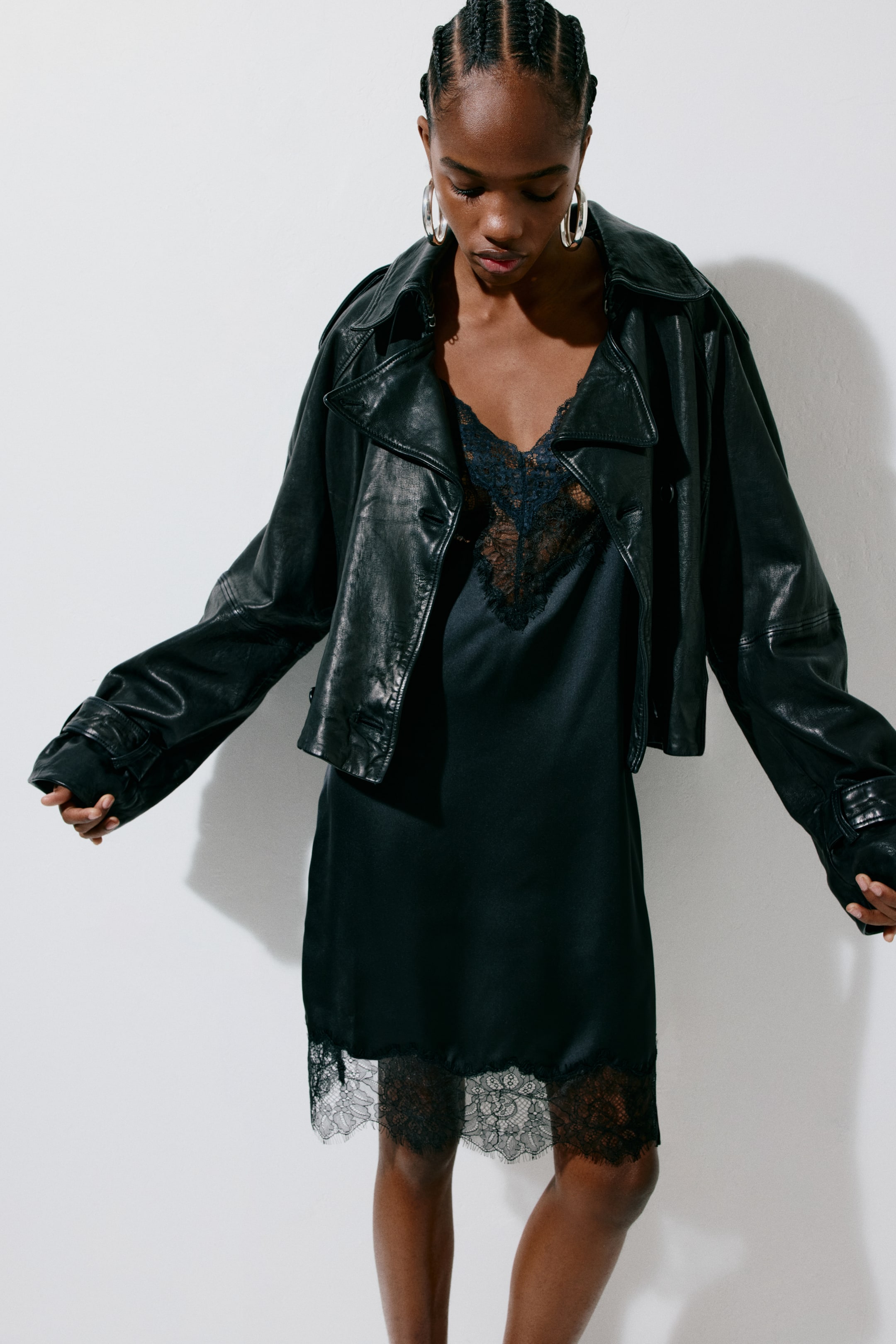 View larger image: A black satin slip dress showcases a V-neckline adorned with black lace trim, complemented by a matching lace-trimmed hem. An oversized black leather jacket is styled open over the dress, highlighting wide lapels and buckled cuffs.