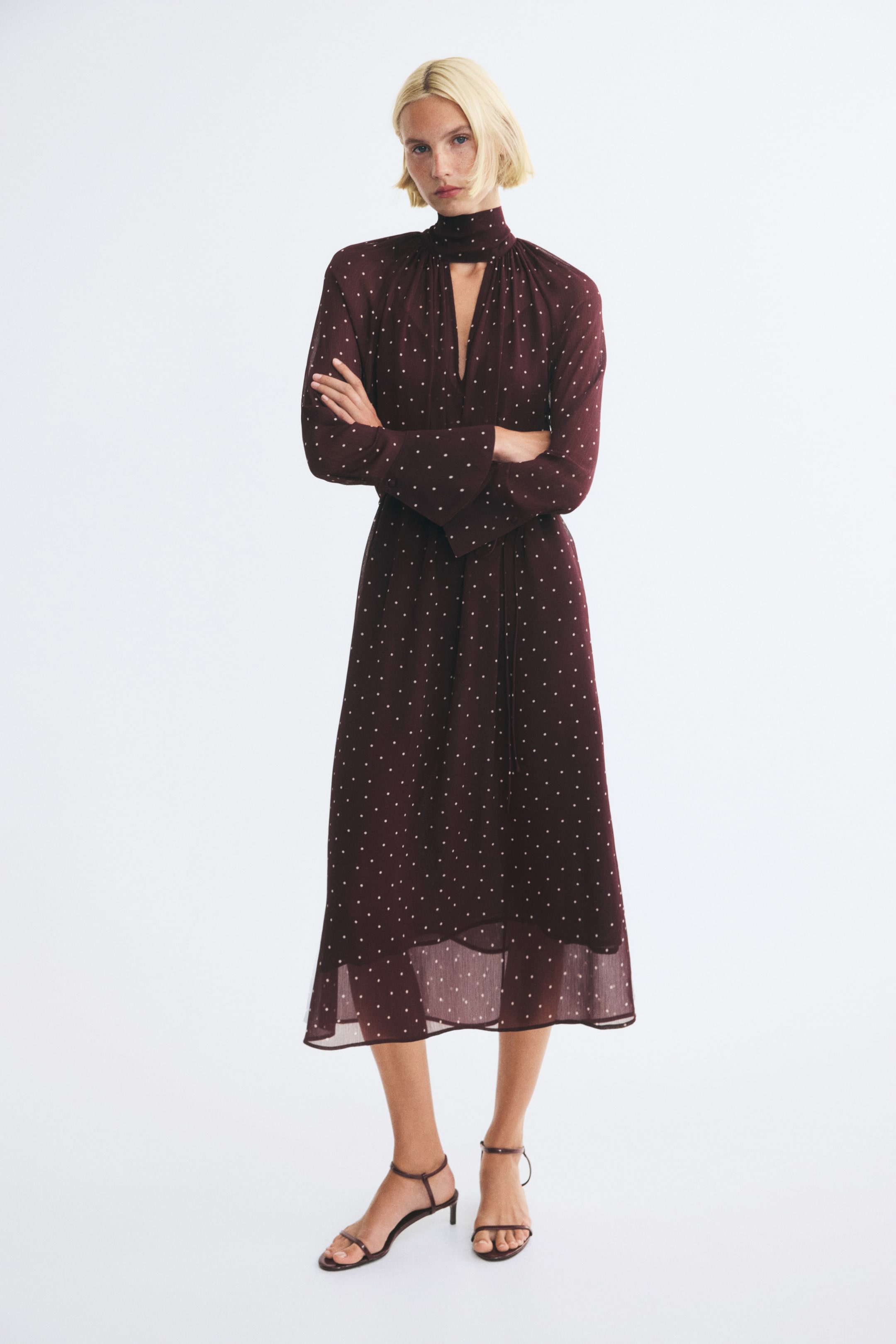 Agrandir l'image: Une robe tunique mi-mollet en bordeaux foncé à motif pois blancs est portée par le mannequin. La robe présente un col montant façon foulard avec une découpe en V sur le devant, des manches raglan longues avec poignets évasés, ainsi qu’une ceinture à nouer assortie à la taille, portée avec des sandales à talons sombres à brides.