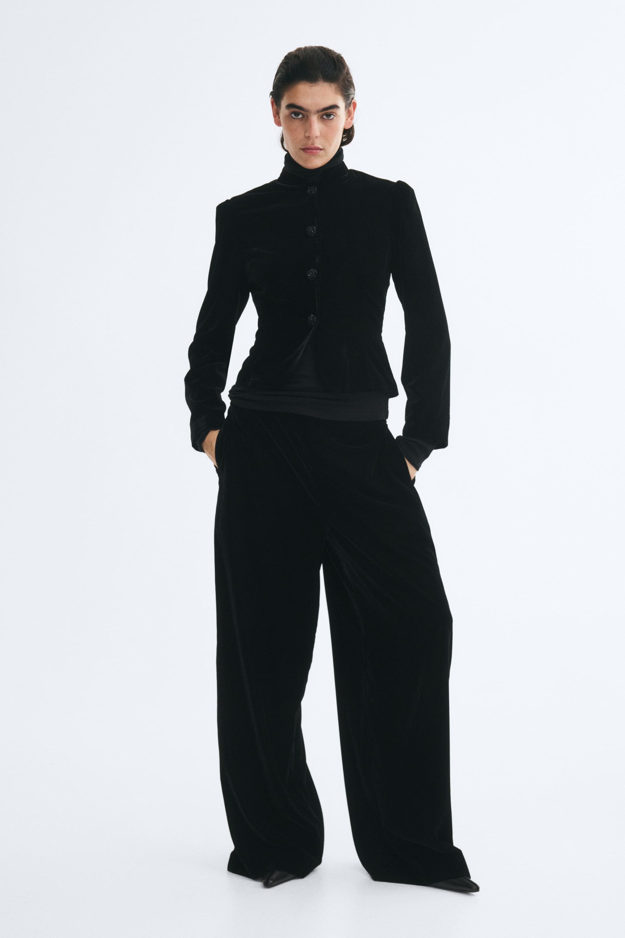 View larger image: Black velvet trousers featuring a long, wide-leg silhouette are worn by the model, who is standing with her hands placed in the side pockets. The trousers drape fully over the feet, partially revealing black pointed-toe shoes, and are styled with a matching black velvet jacket.