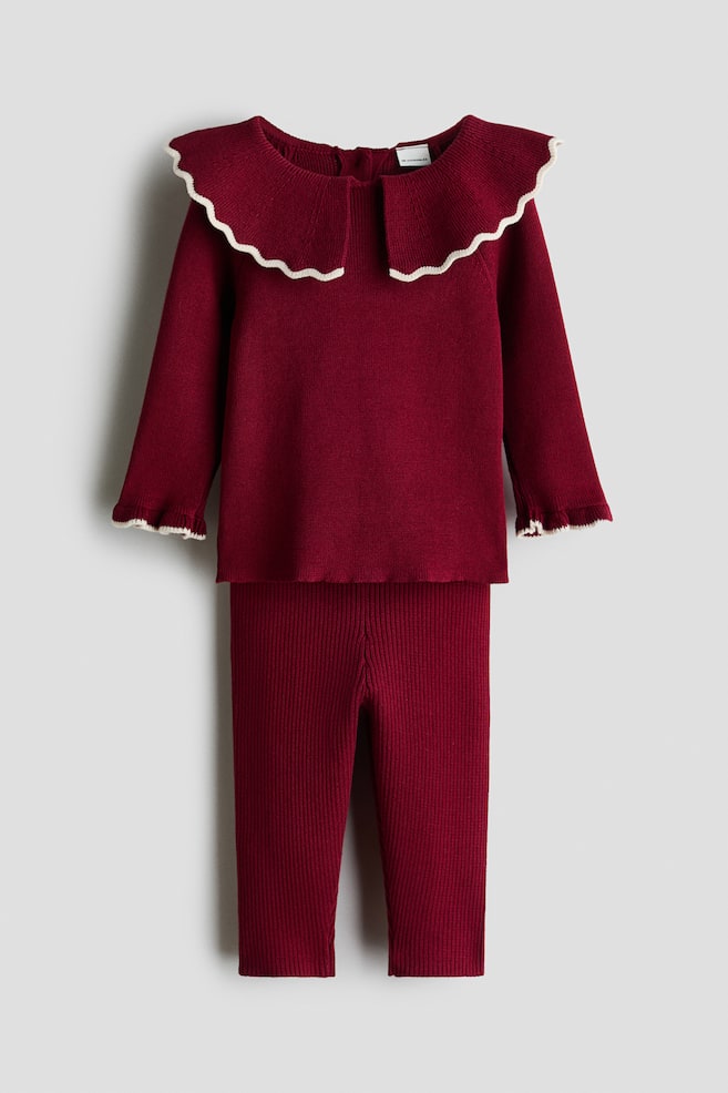 A front-facing view shows a dark red knitted two-piece set comprising a long-sleeved jumper and matching ribbed leggings. The jumper features a large scalloped collar and frilled cuffs, both detailed with a contrasting cream wavy trim. - 1
