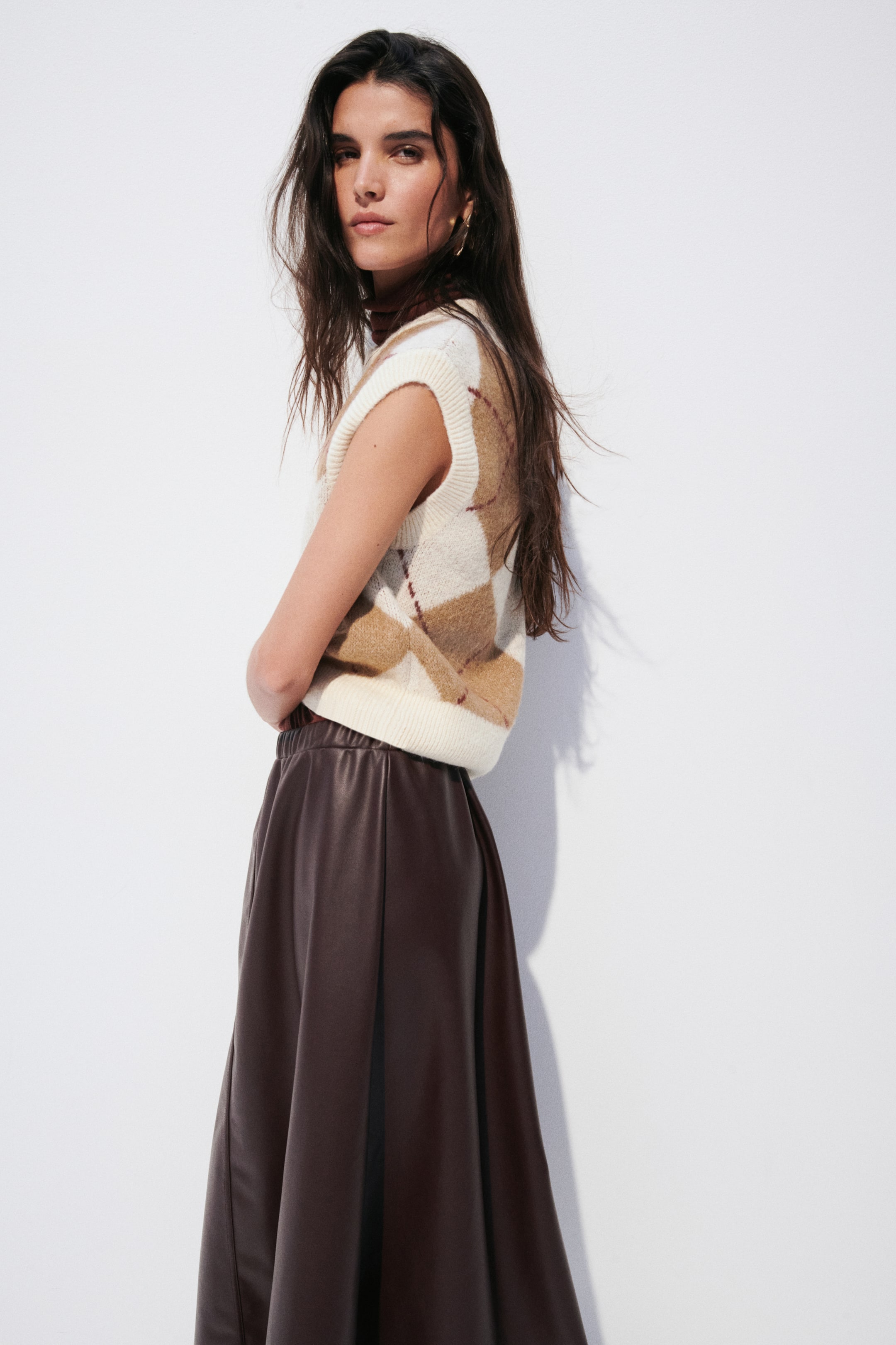 View larger image: A sleeveless jumper vest is being worn, showcasing a cream base with a brown and dark red argyle pattern, ribbed armholes, and a ribbed hem, layered over a dark brown top. A dark brown faux leather skirt with an elasticated waistband is also being worn, flowing in an A-line silhouette.