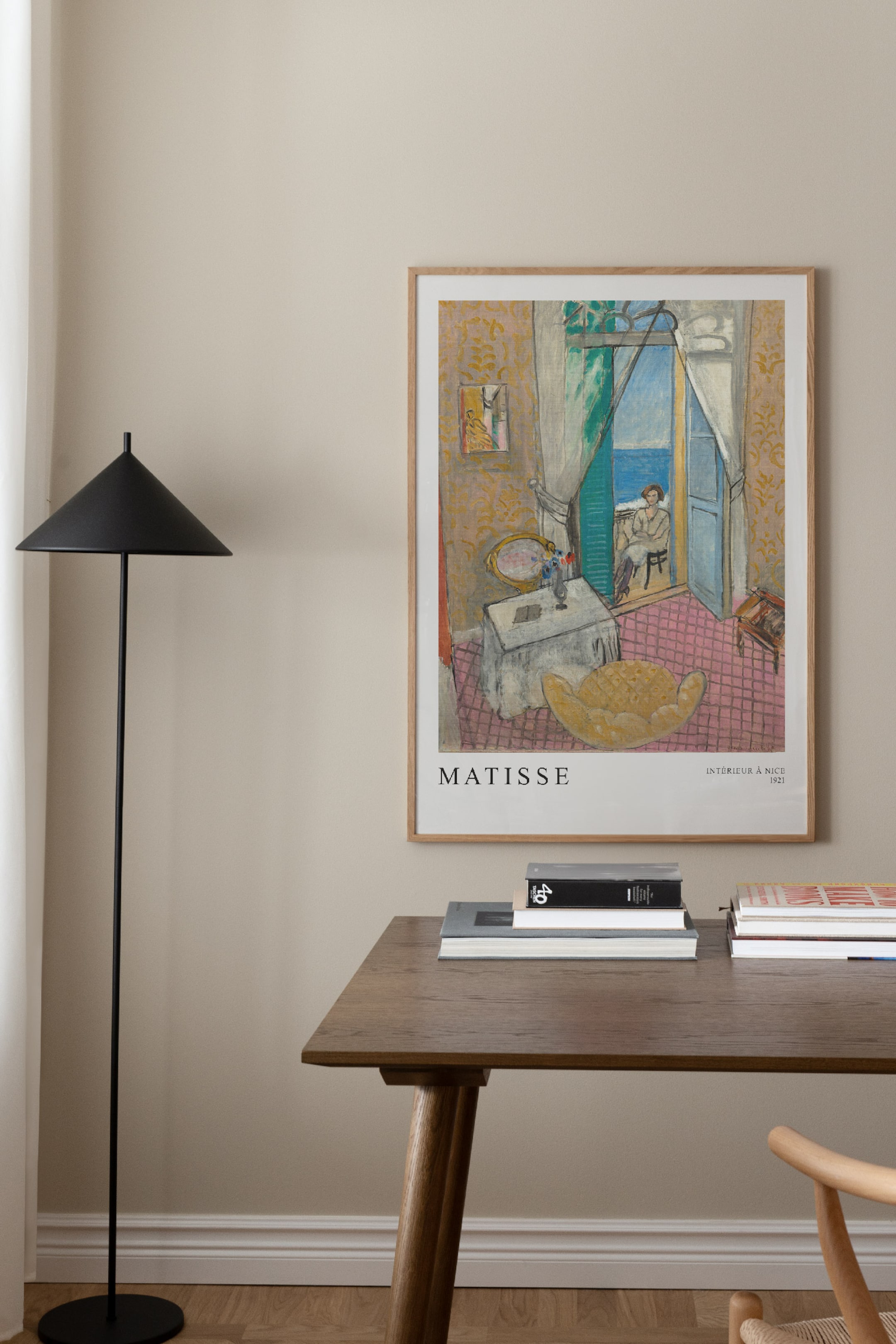 Grotere afbeelding bekijken: Een ingelijste poster hangt verticaal en toont Matisse's kunstwerk 'Intérieur à Nice 1921'. Het kunstwerk beeldt een interieur uit met een vrouw bij een raam met uitzicht op zee en is omlijst met een dunne, lichtbruine houten lijst.