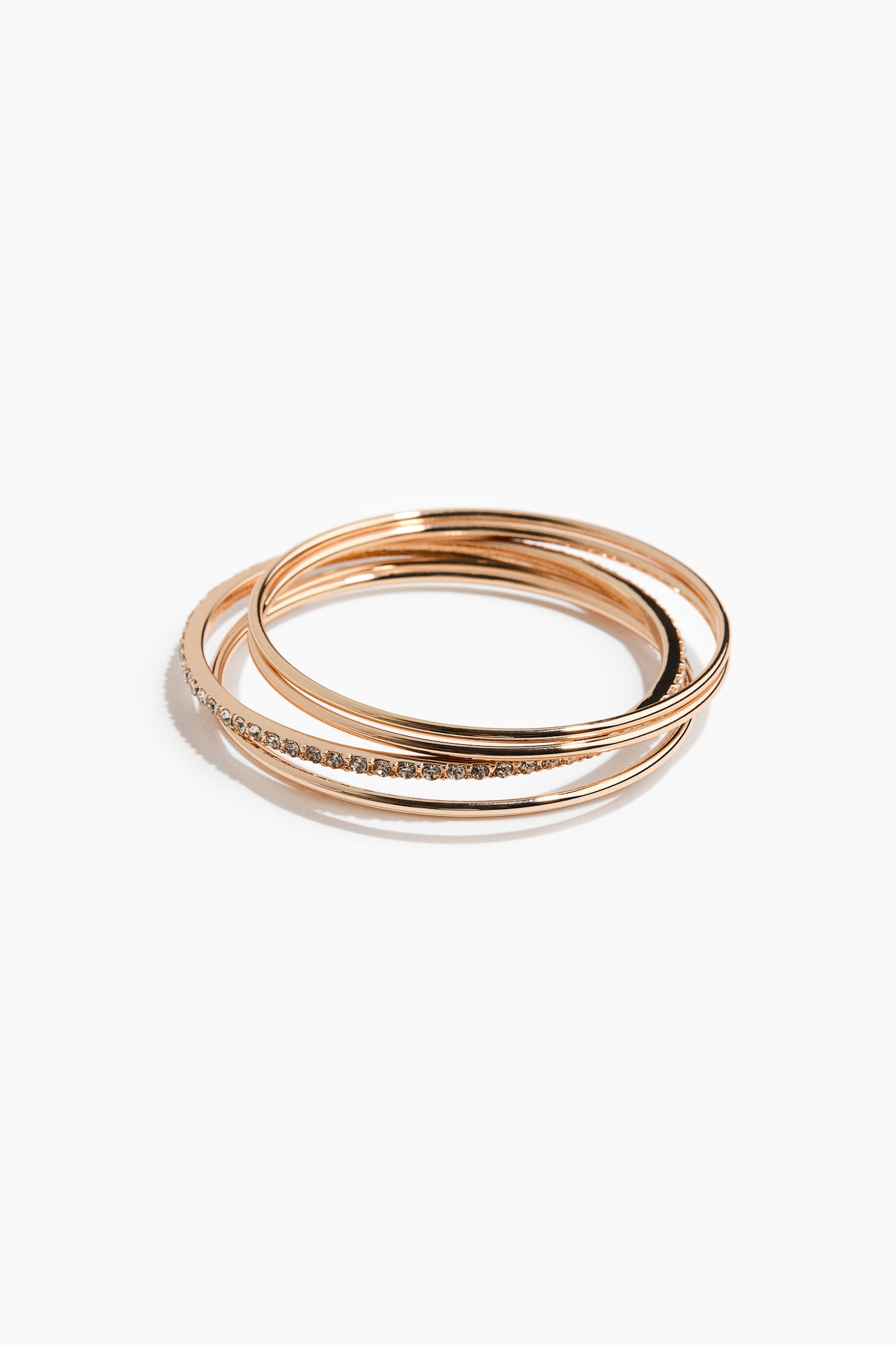 Un conjunto de pulseras rÃ­gidas de tono dorado reposa en una pila superpuesta, con una de las bandas adornada con una hilera de pequeÃ±os cristales transparentes y tres bandas lisas y pulidas.